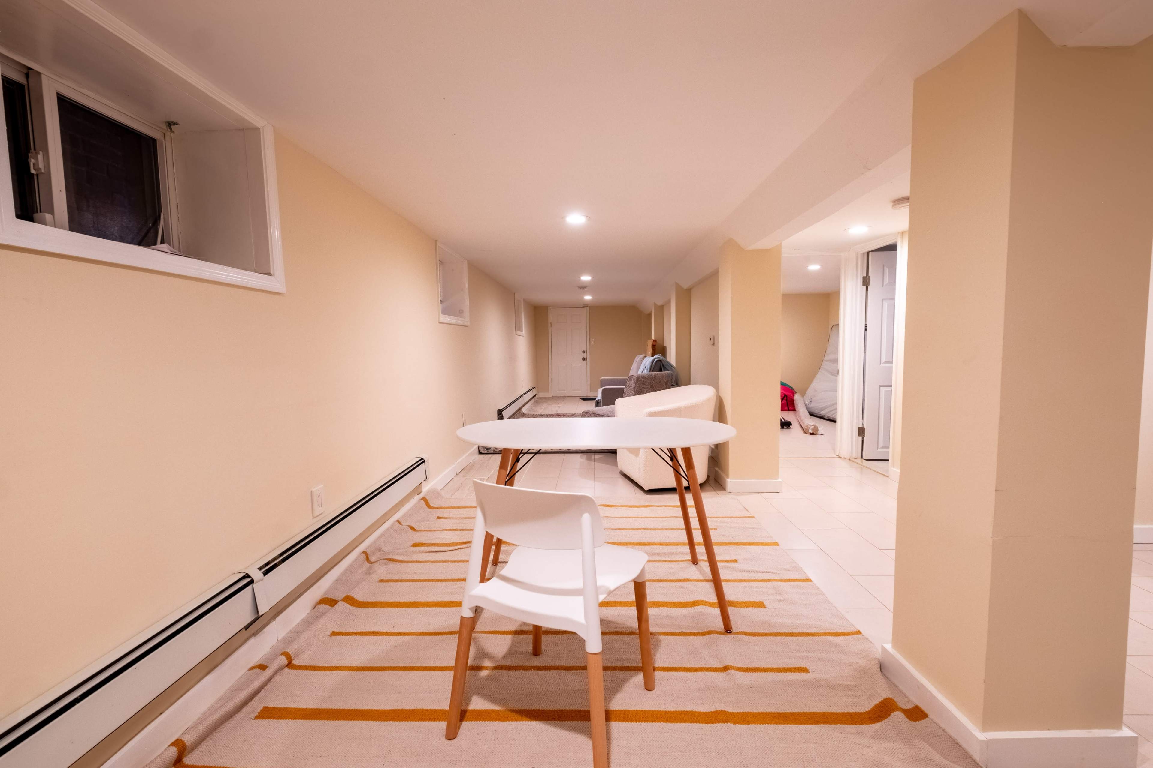 A narrow, softly lit basement room with a table and two chairs on a striped rug, along with a couch and a closed door in the background.