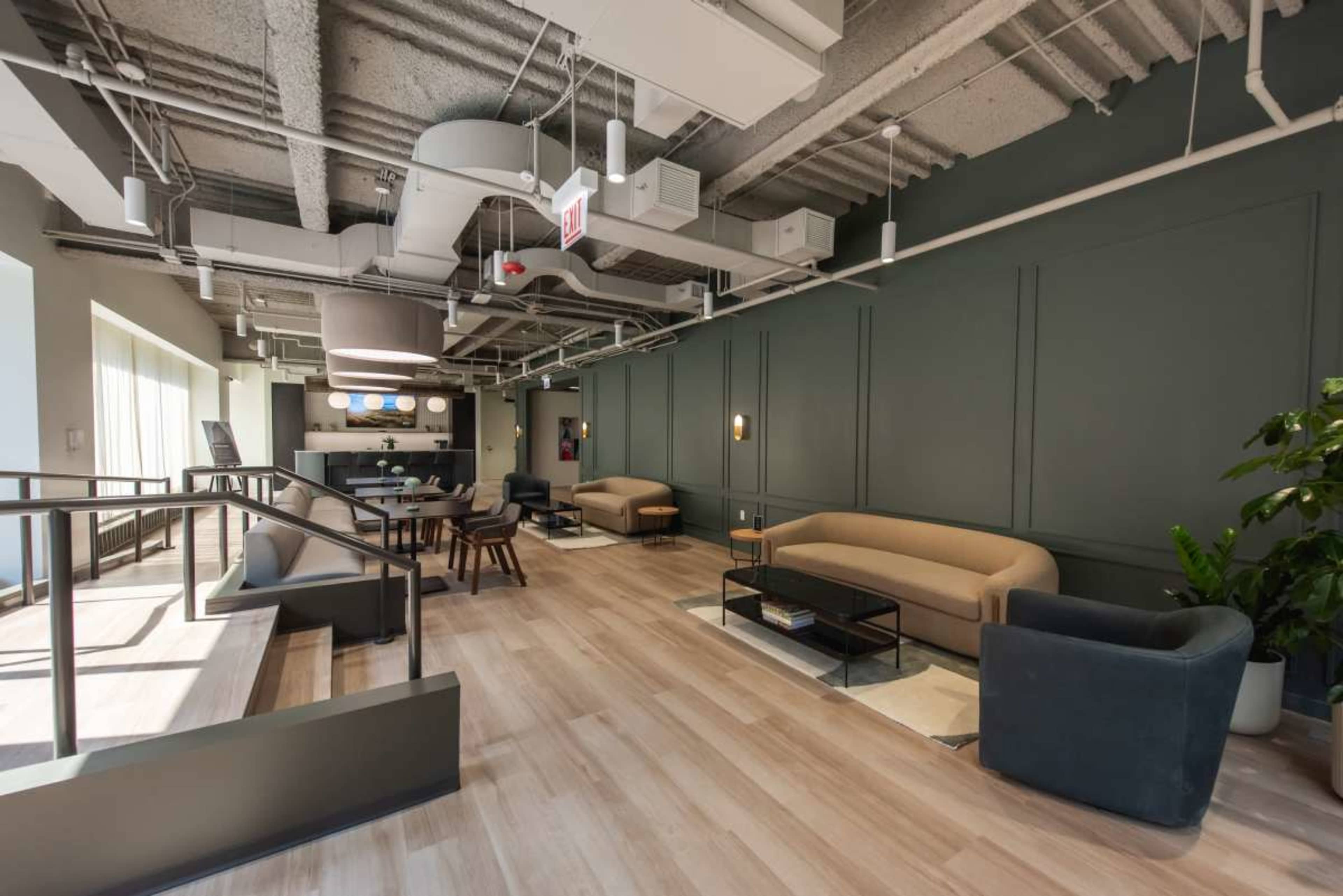 The image shows a modern interior lounge area with light wood flooring, dark green walls, and various seating arrangements including couches and chairs.