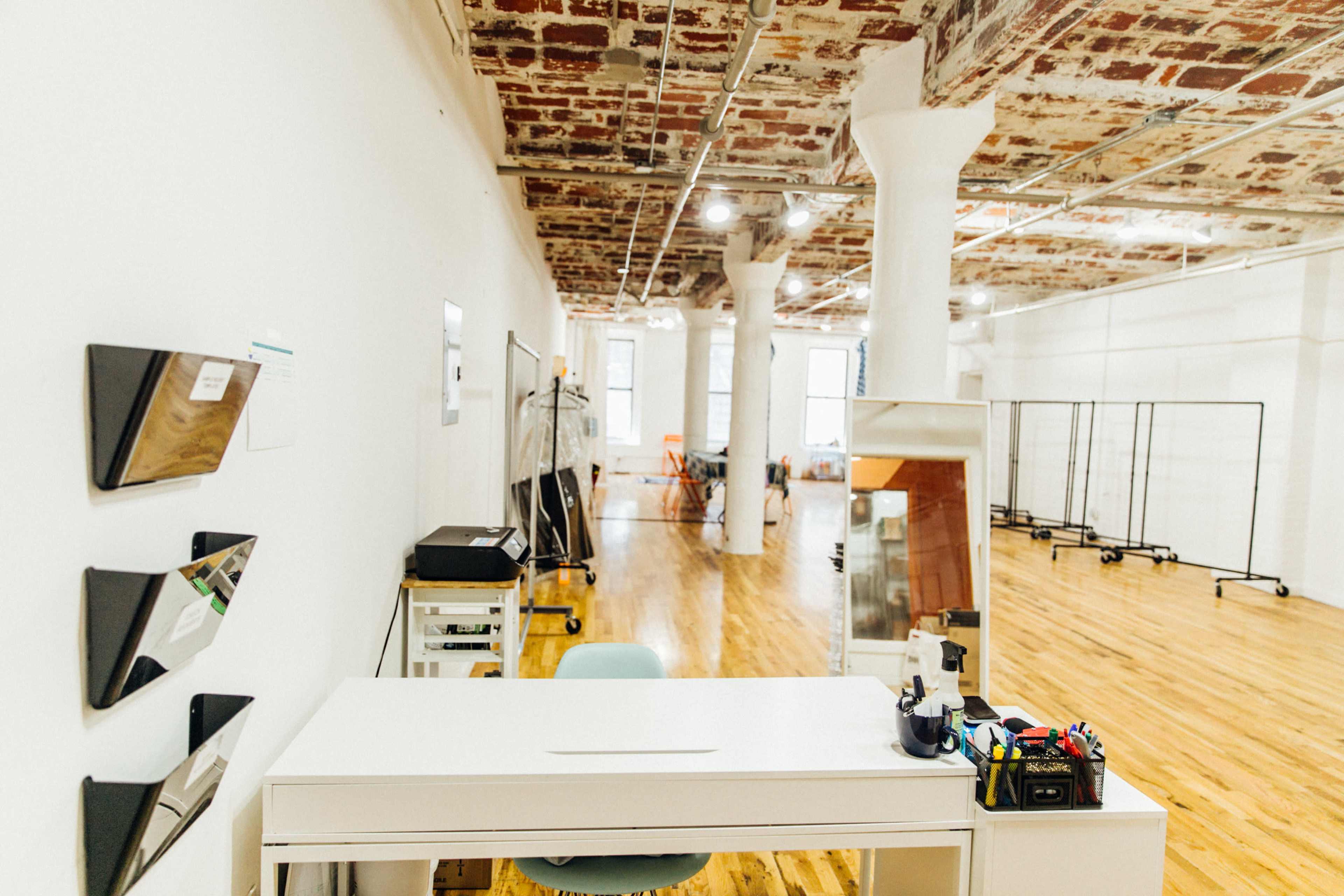 A bright, open workspace with a white desk, wooden flooring, and exposed brick ceilings, featuring minimal furnishings and studio equipment in the background.