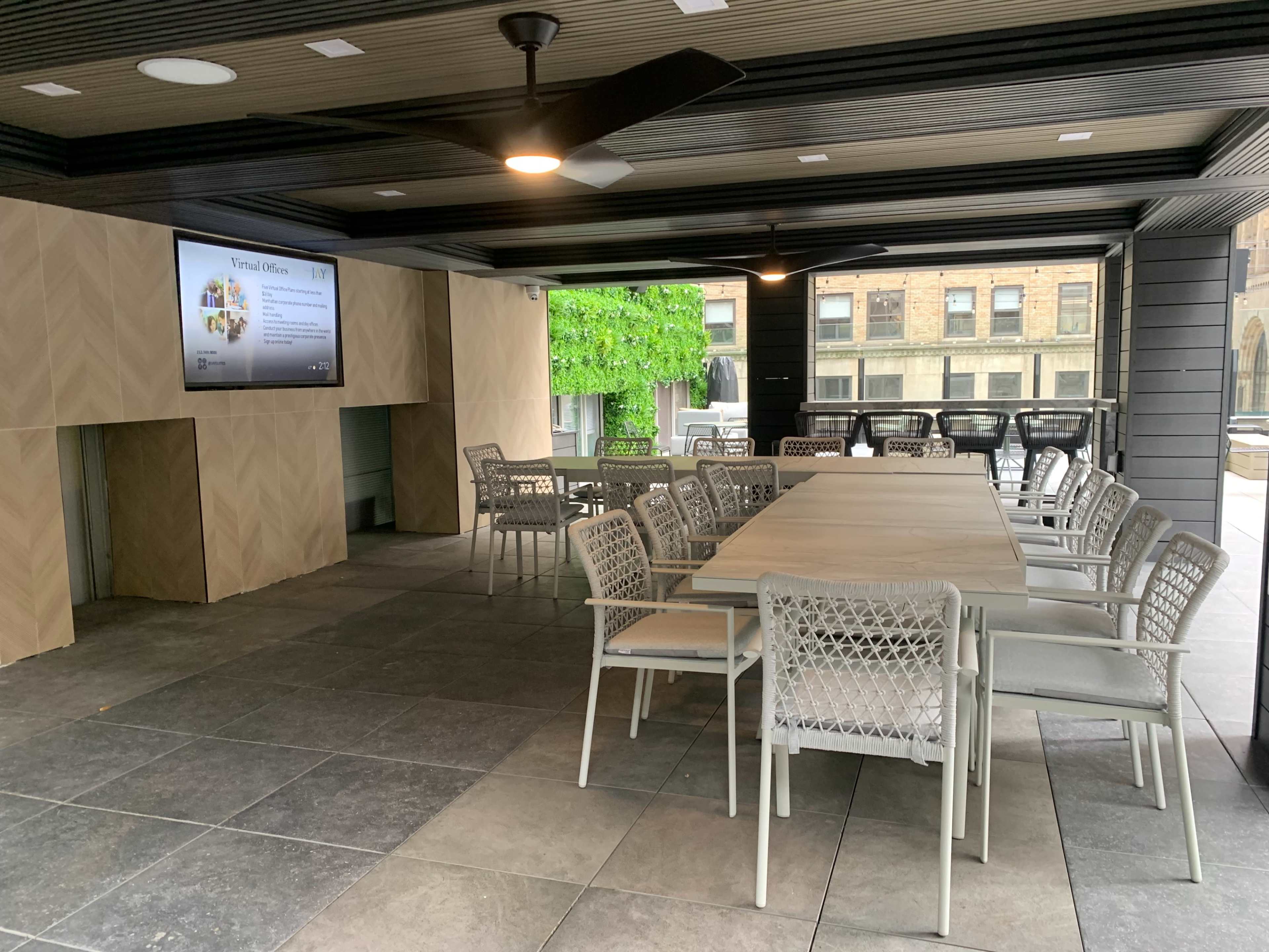 The image shows a covered outdoor space featuring a long table with seating, a presentation screen, and green foliage in the background.
