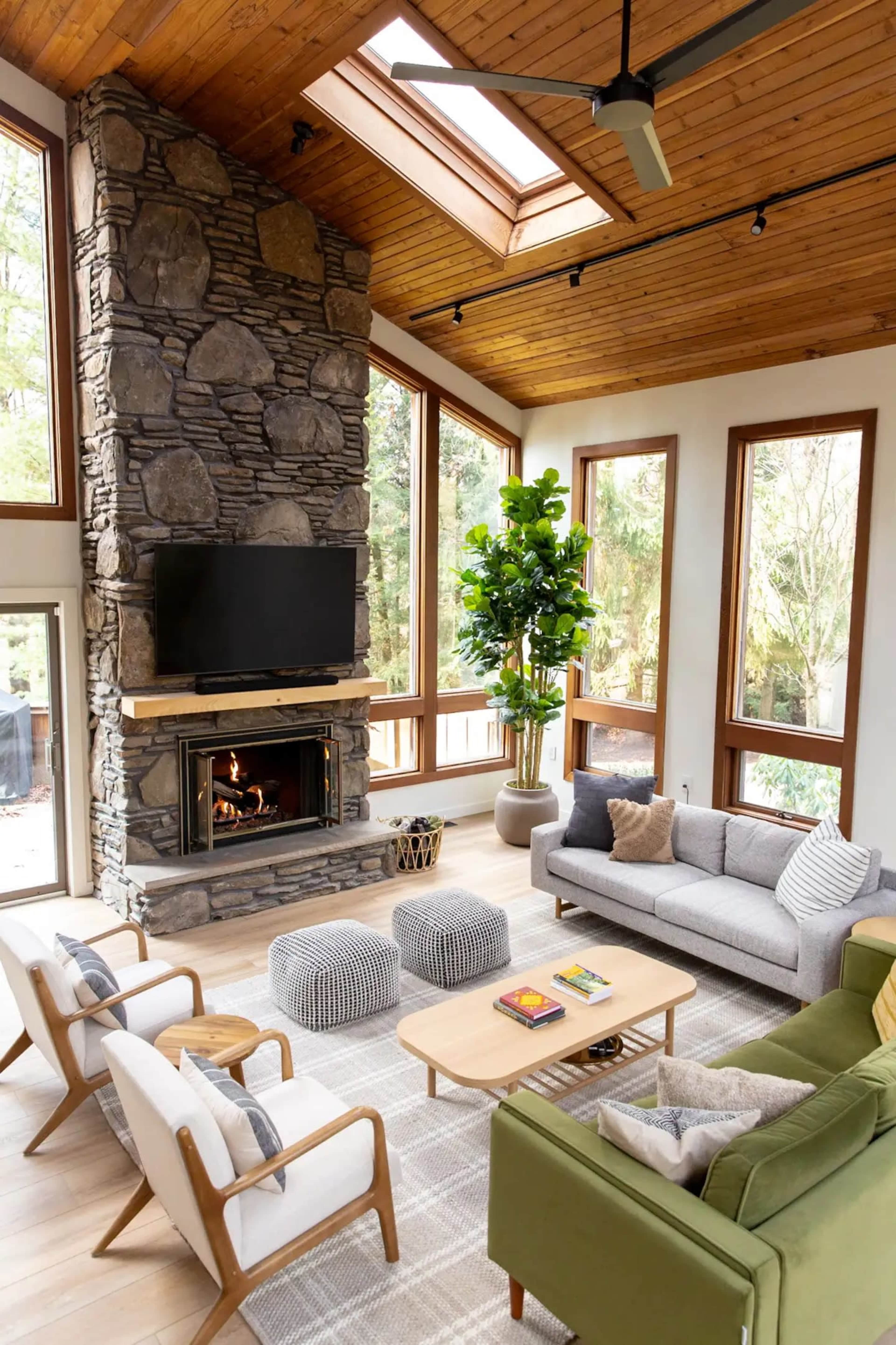 The living room features a stone fireplace with a mounted television, surrounded by large windows that allow natural light to fill the space, along with a mix of modern furniture including a green sofa and wooden accent tables.