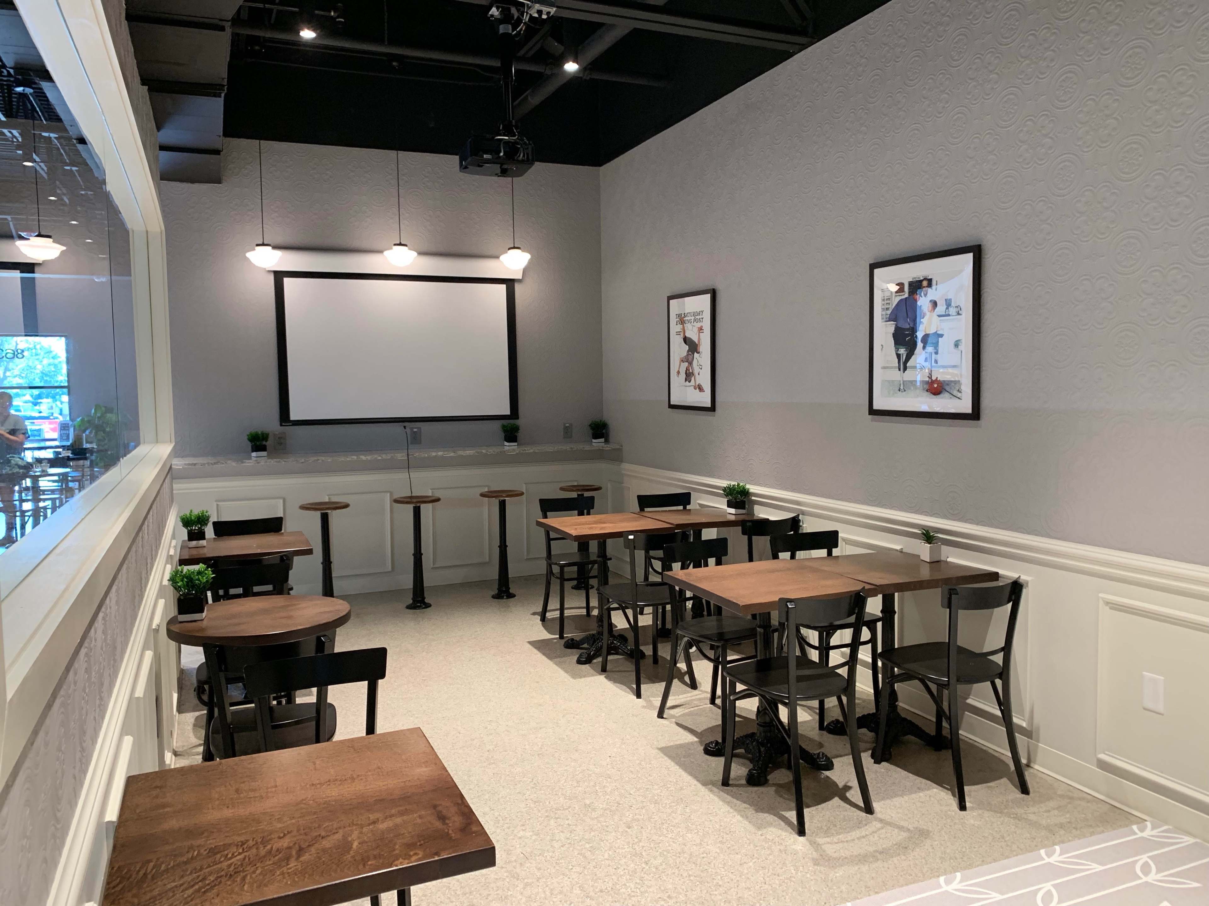 A modern café interior with several tables and stools arranged against a light gray wall featuring a projector screen and framed artwork.