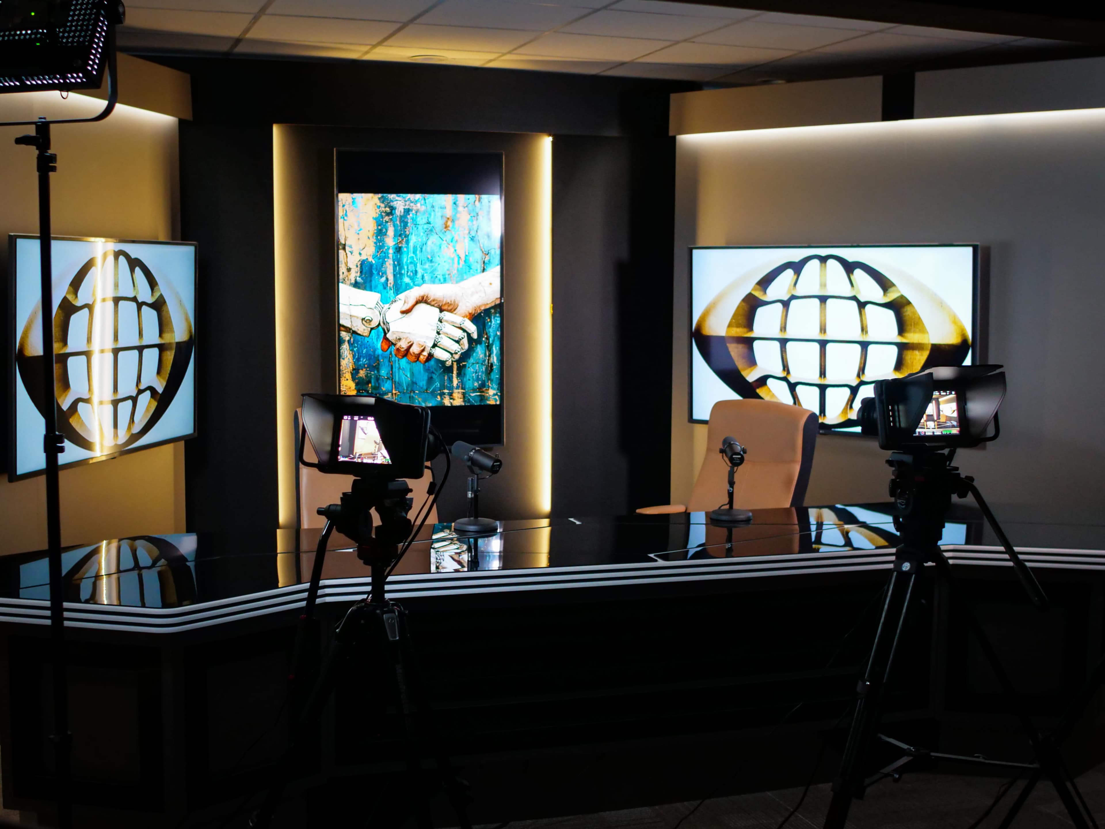 A television studio setup features a central desk with cameras and monitors displaying handshake artwork against a backdrop of warm lighting.