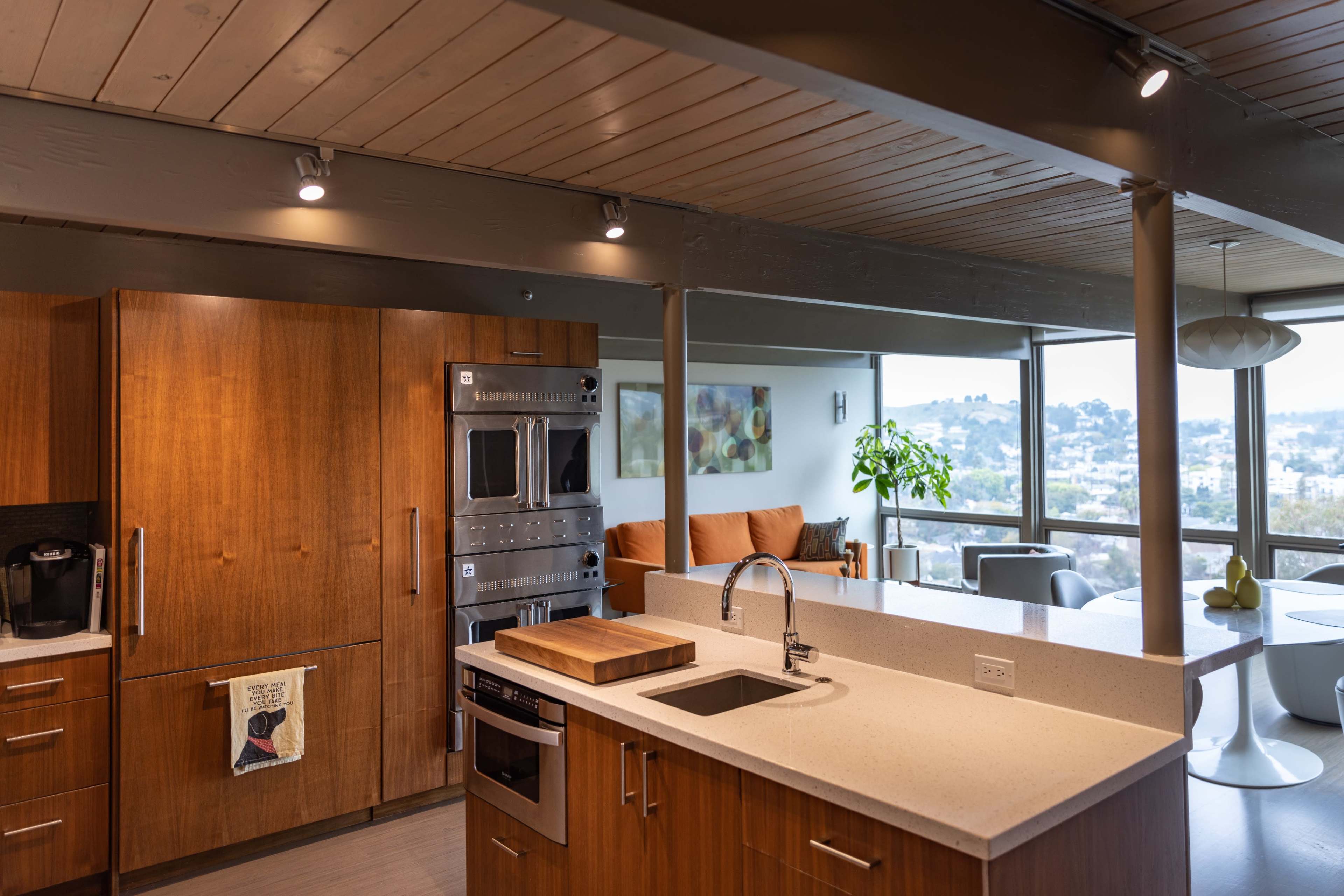 A modern kitchen features wooden cabinetry, stainless steel appliances, and a view of a city skyline through large windows.