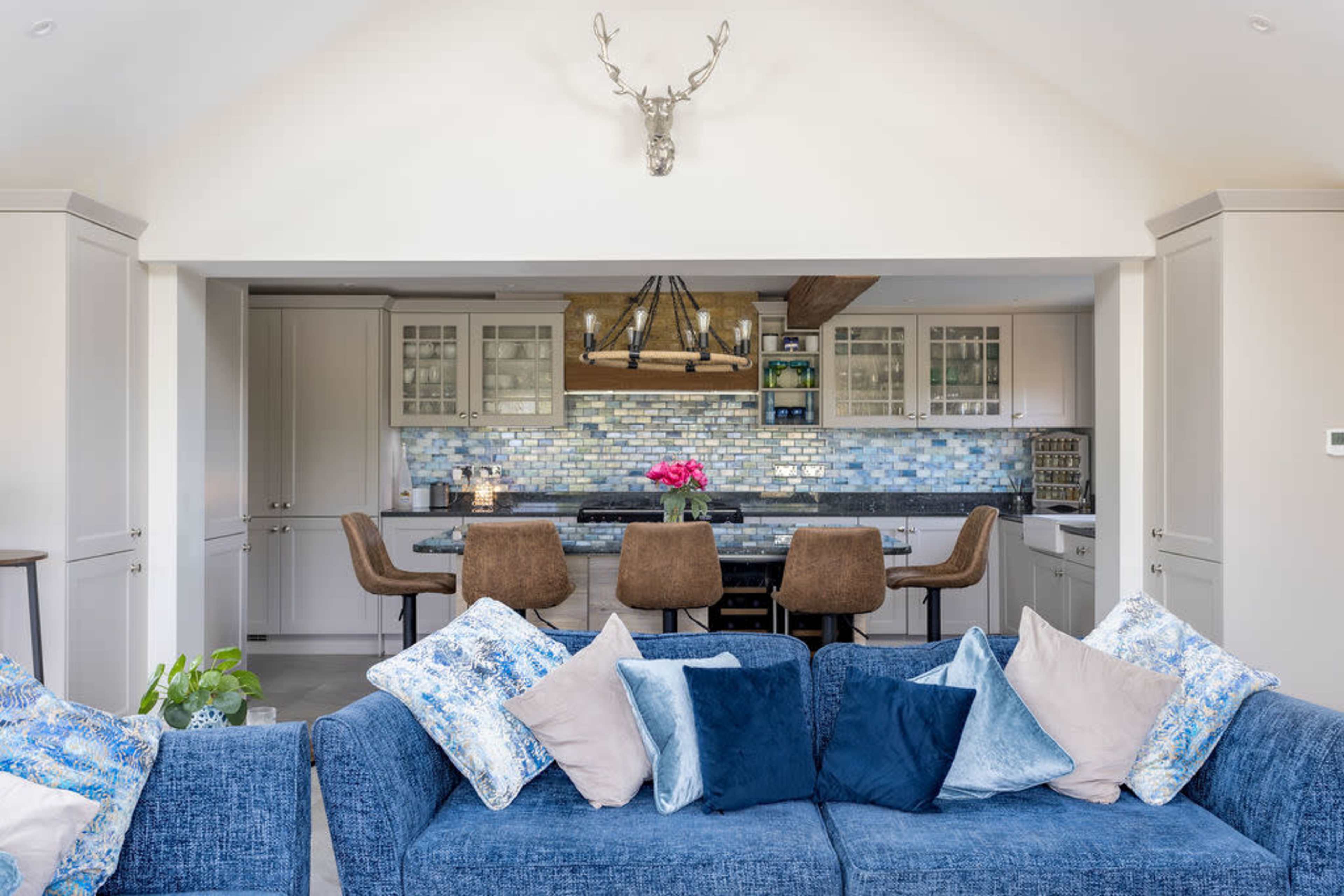 A modern kitchen with a blue mosaic backsplash and a dining area featuring bar stools is visible from a living space with a blue sofa decorated with various cushions.