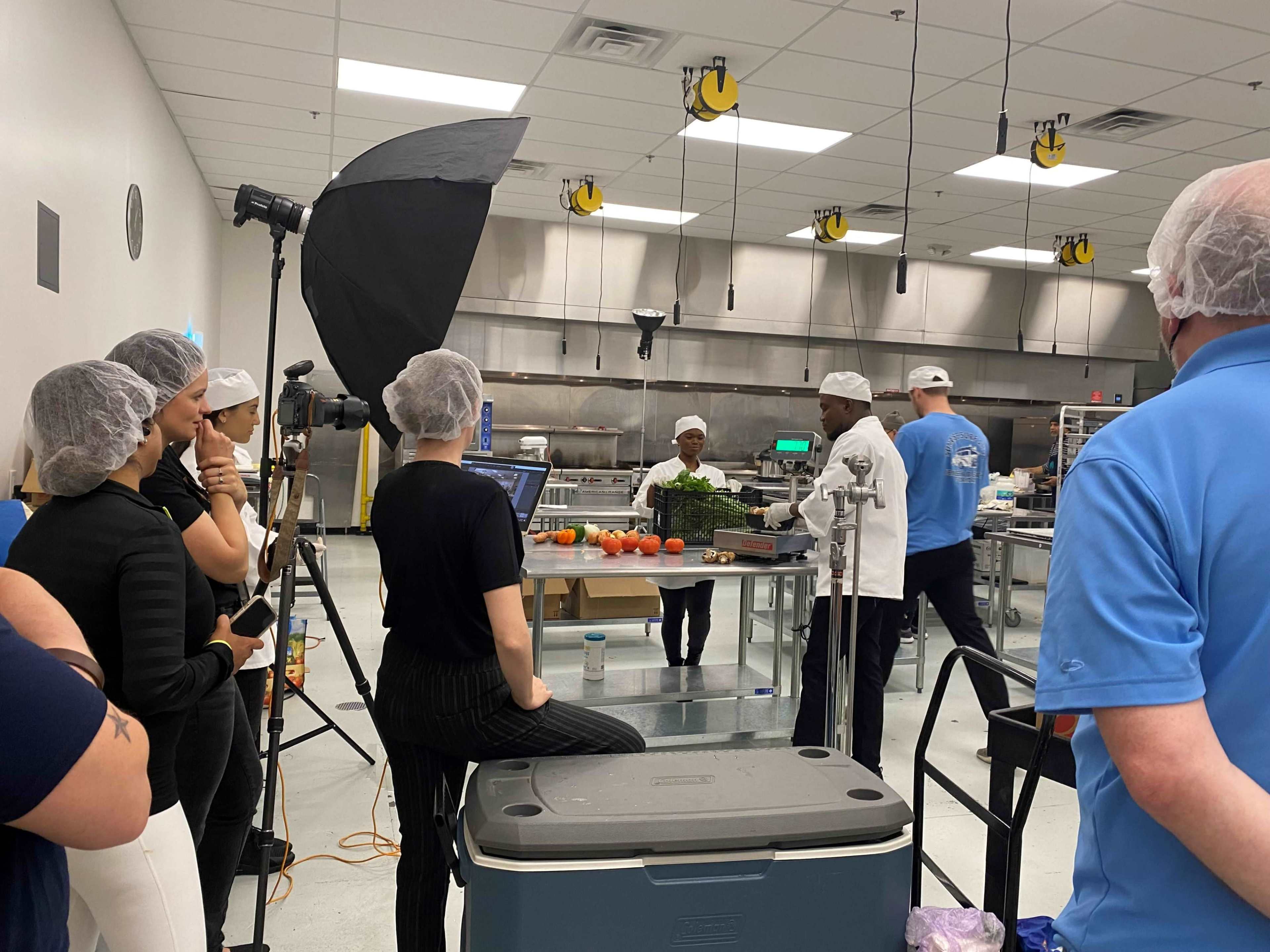 A group of people observes a cooking demonstration in a commercial kitchen setting.