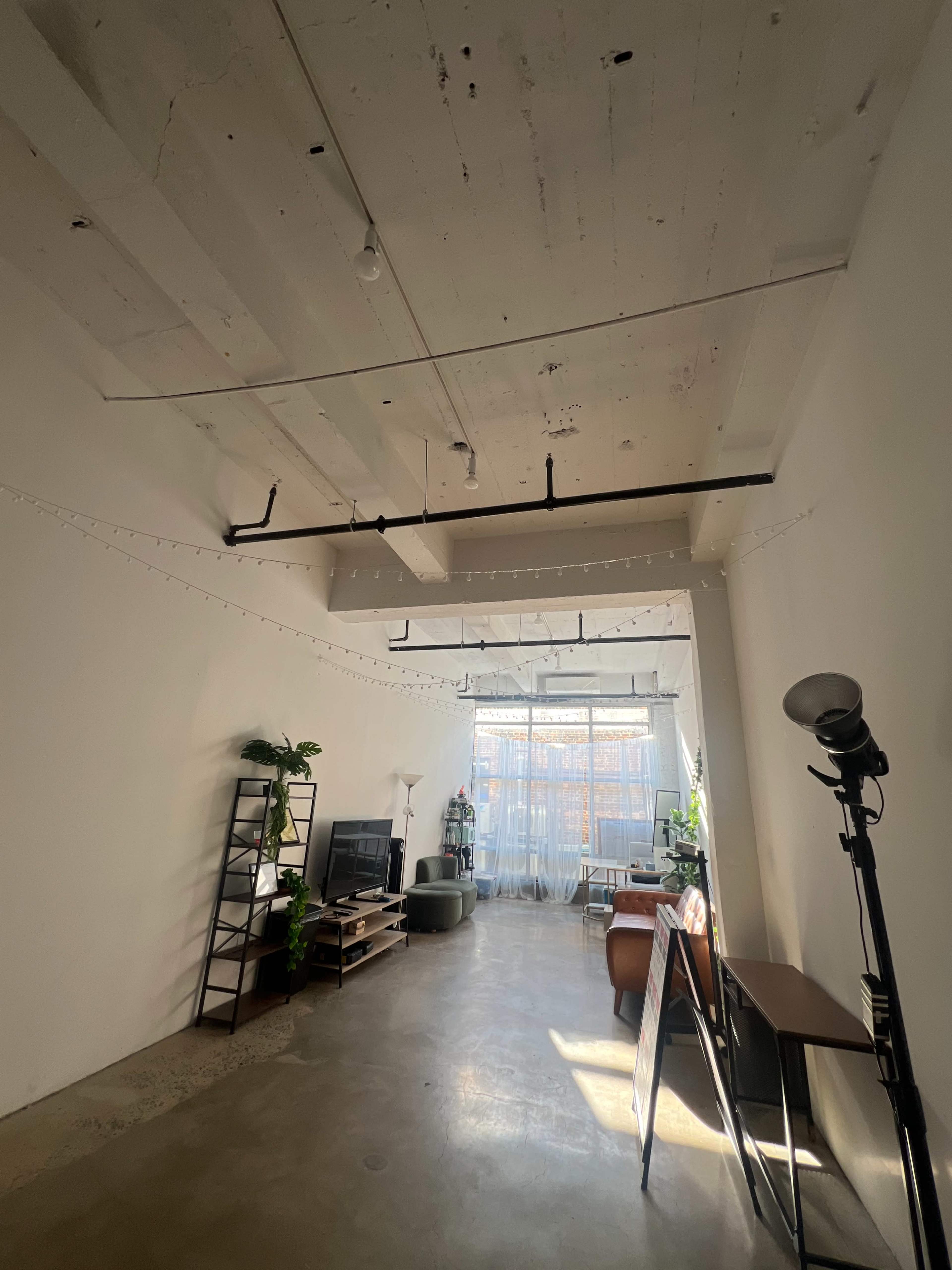 The image shows an industrial-style room with concrete floors, exposed ceiling beams, and large windows letting in natural light, filled with plants and minimalist furniture.