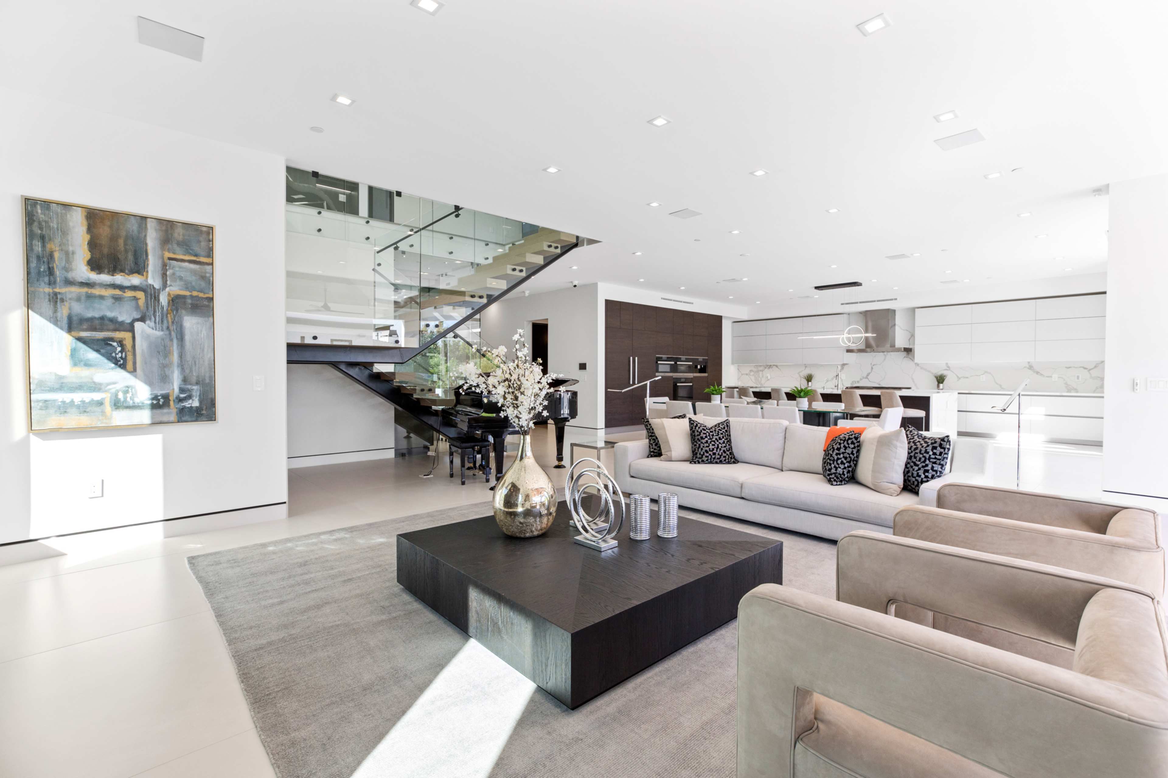 The image shows a modern living room with a large gray sofa, a central coffee table, and a glass staircase leading to an upper level, surrounded by minimalist decor and bright lighting.