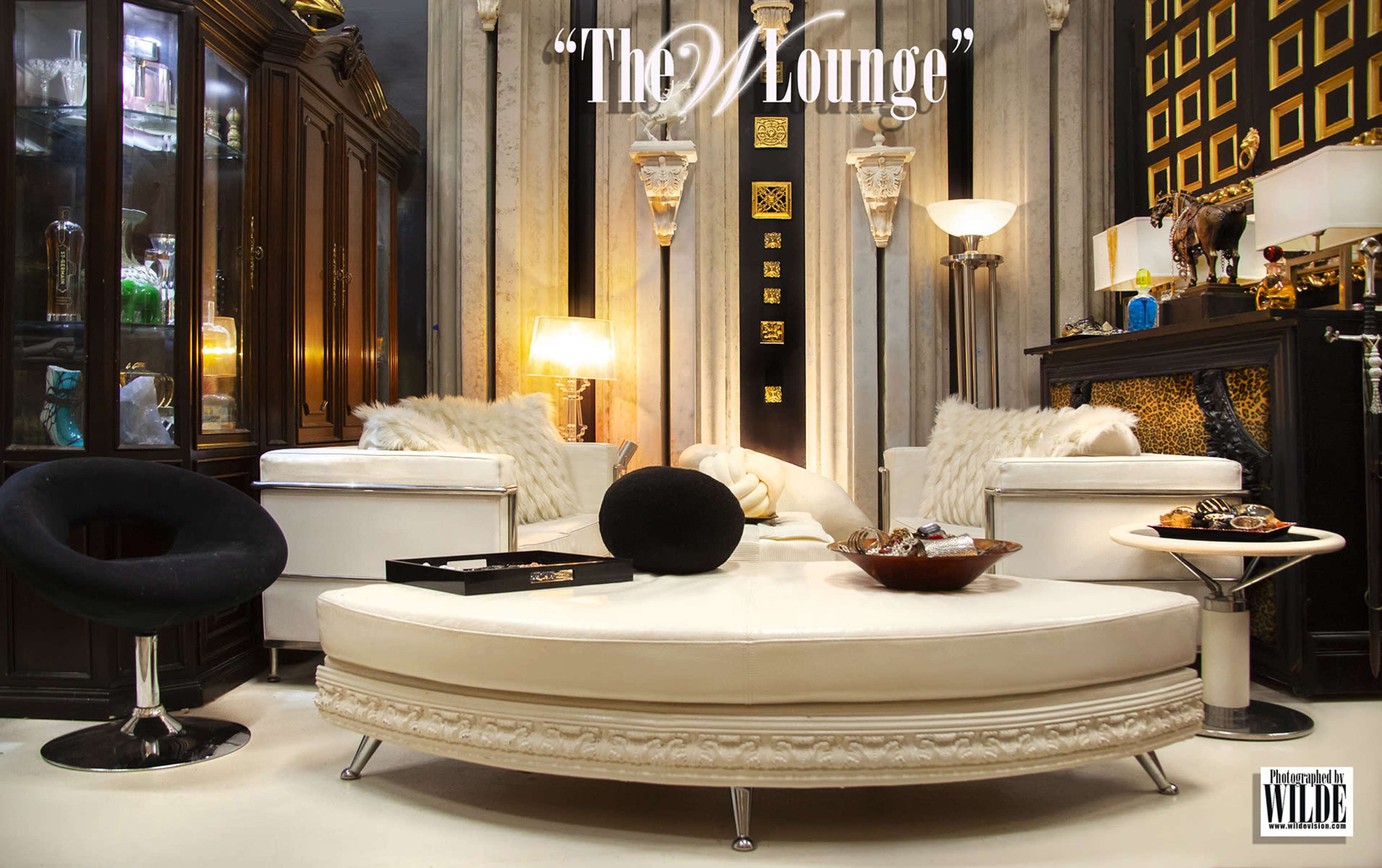 The image features a stylishly decorated lounge area with a curved white sofa, two black chairs, and a glass display cabinet showcasing various decorative items.