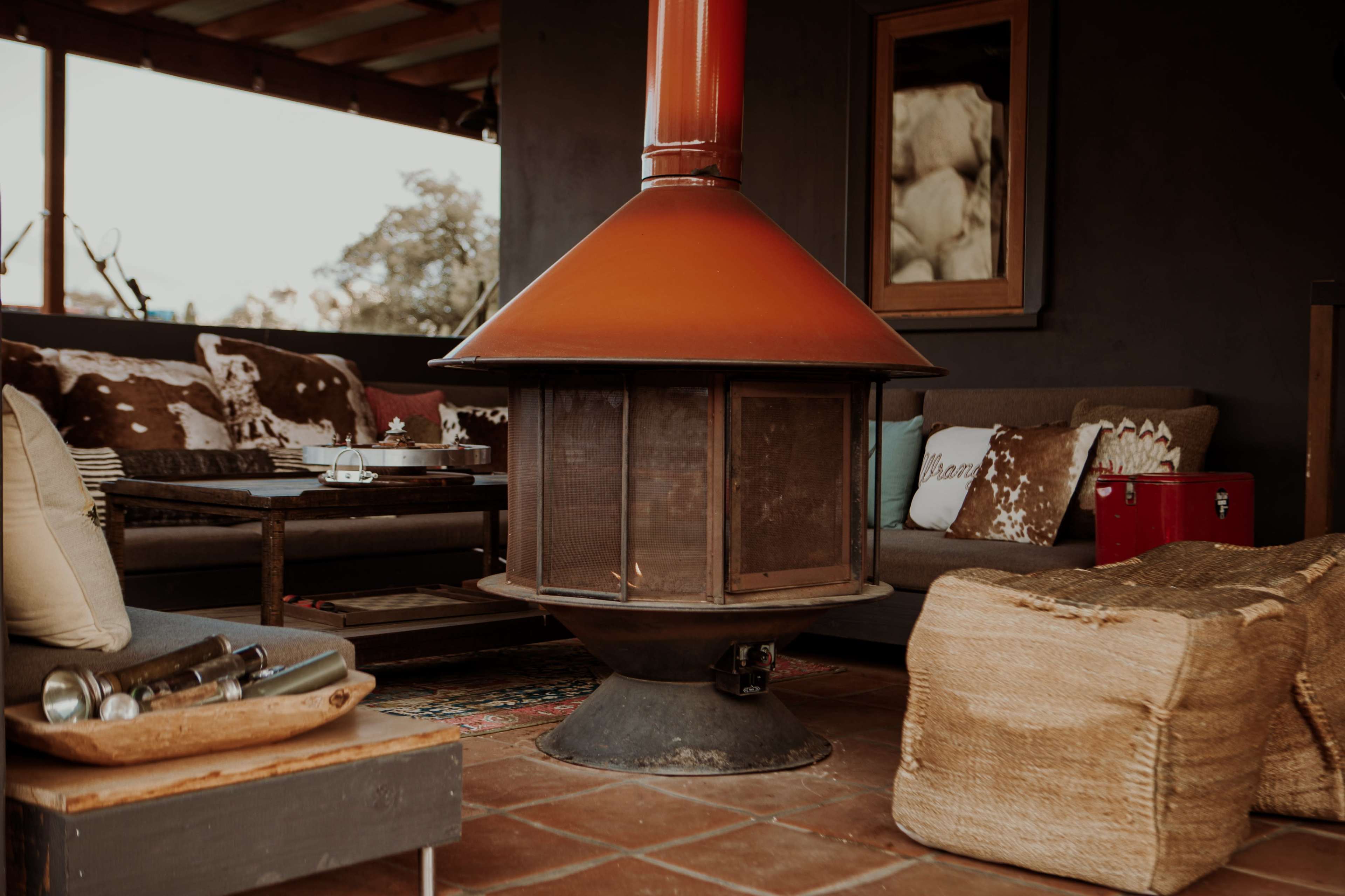 The image shows a cozy living area featuring a central circular wood-burning stove with a red chimney, surrounded by comfortable seating and rustic decor.