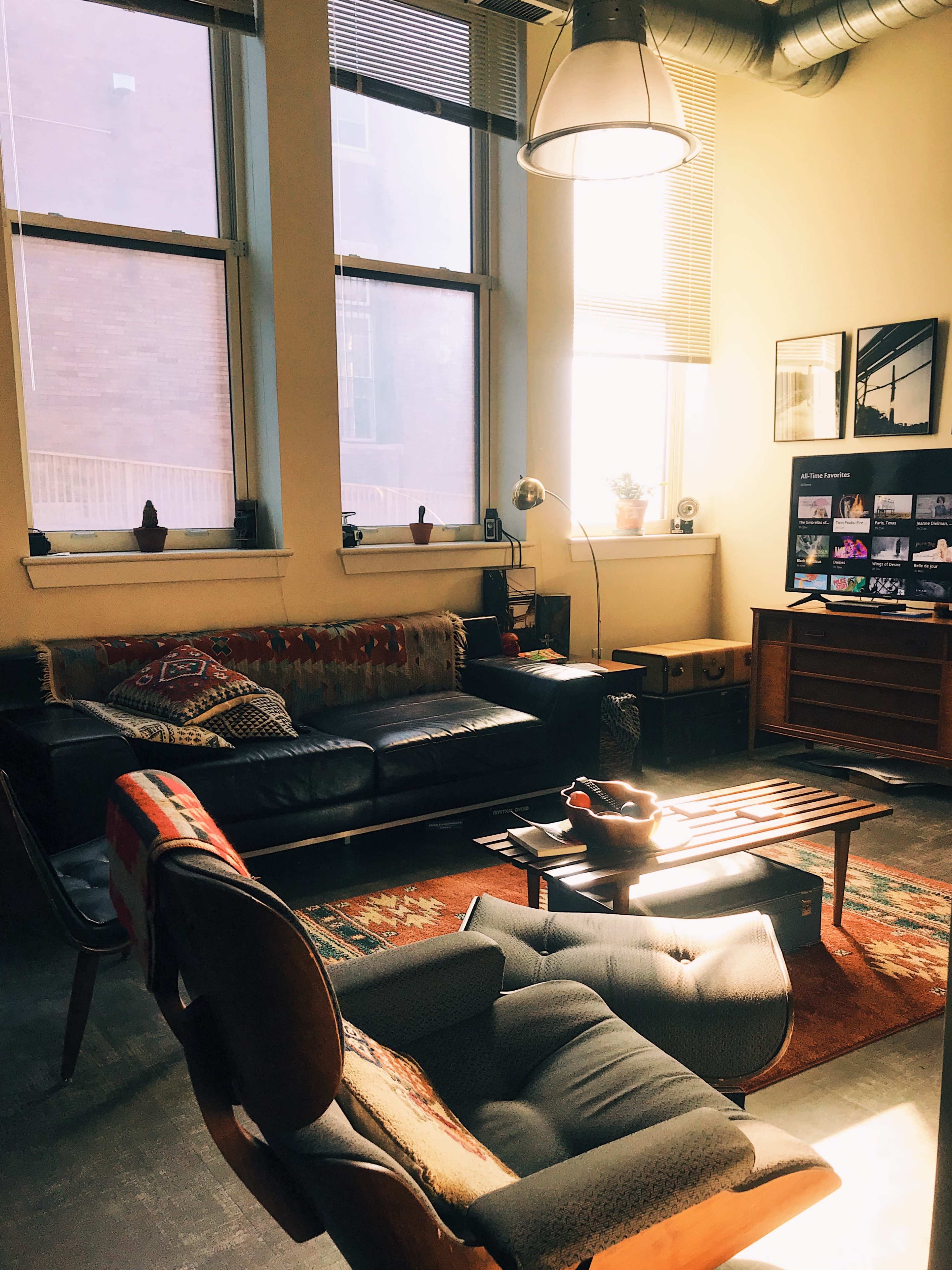 The living room features a leather sofa, a wooden coffee table, an armchair, and large windows allowing natural light to stream in.