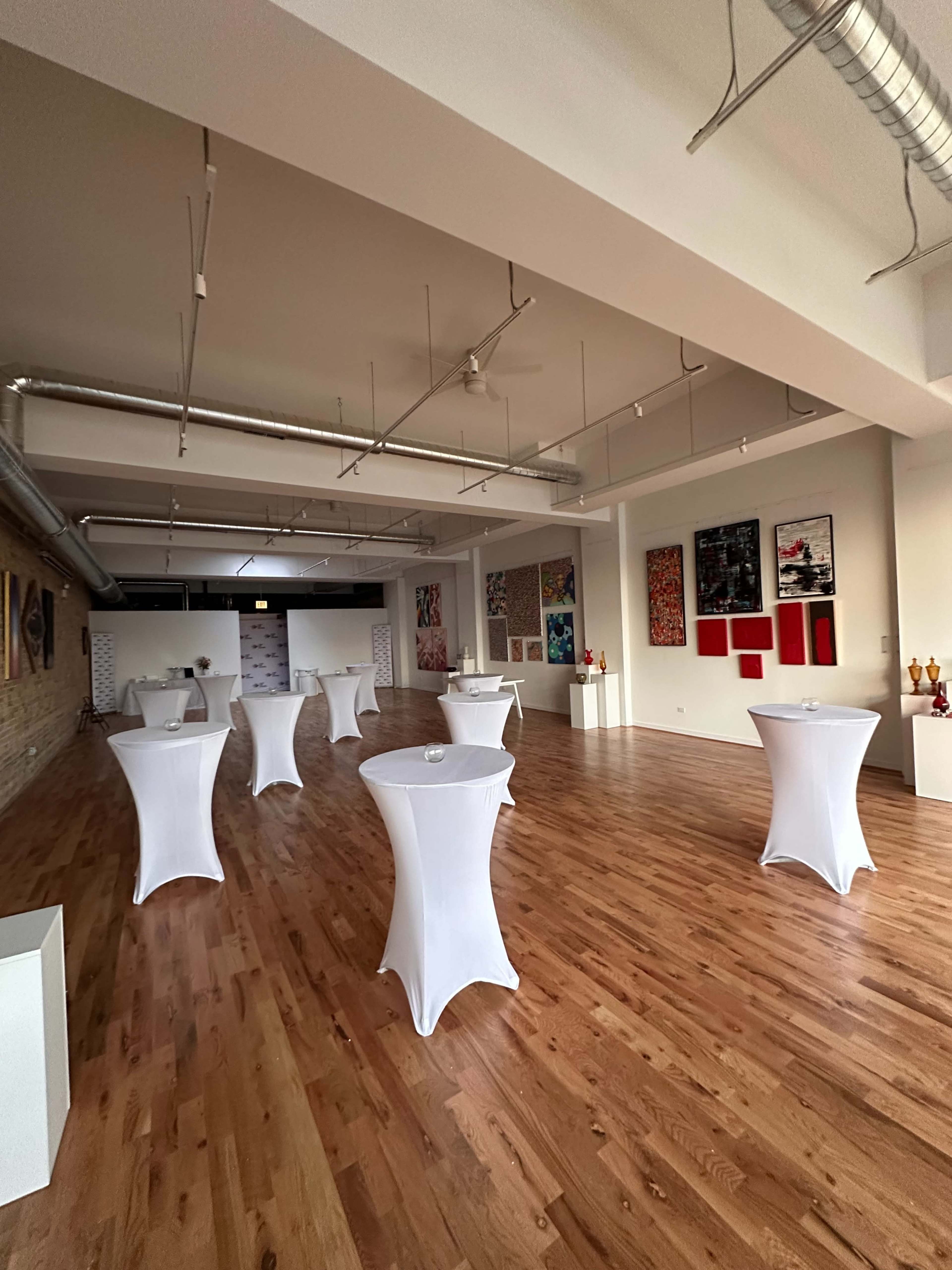 The image shows a spacious interior with wooden flooring, featuring several cocktail tables covered in white linens and various artworks displayed on the walls.