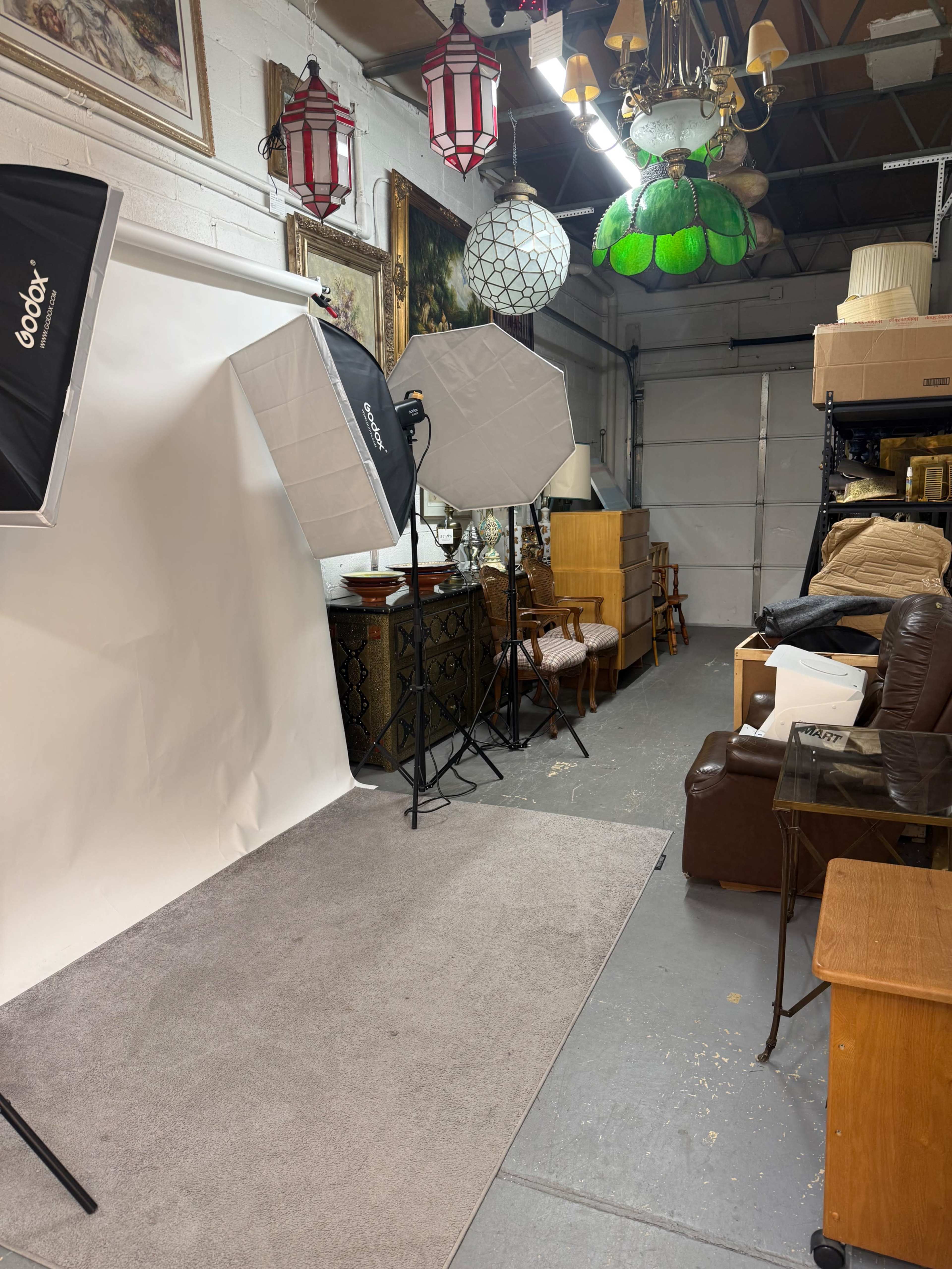 The image shows a photo studio setup in a garage-like space, featuring lighting equipment, a white backdrop, and various furniture items stacked against the walls.