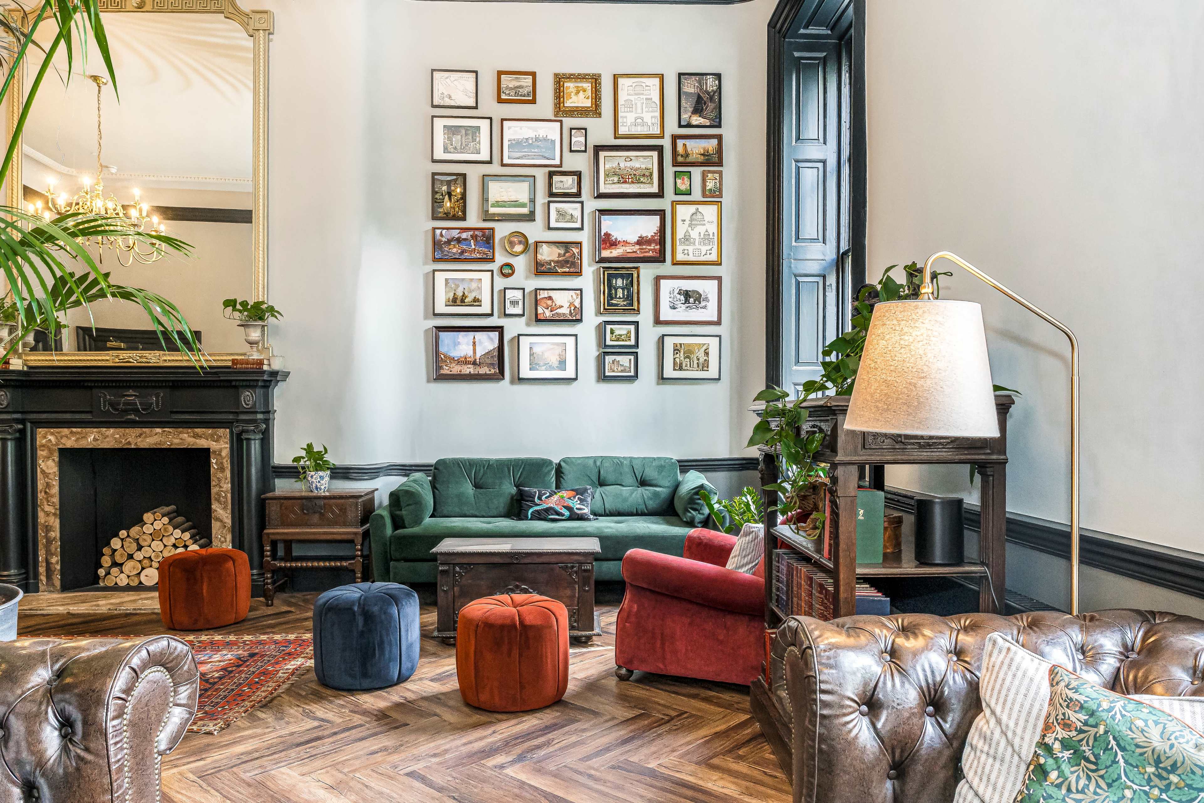 The image shows a stylish living room with a green sofa, various framed artworks on the wall, and a cozy fireplace surrounded by plush furniture and colorful ottomans.