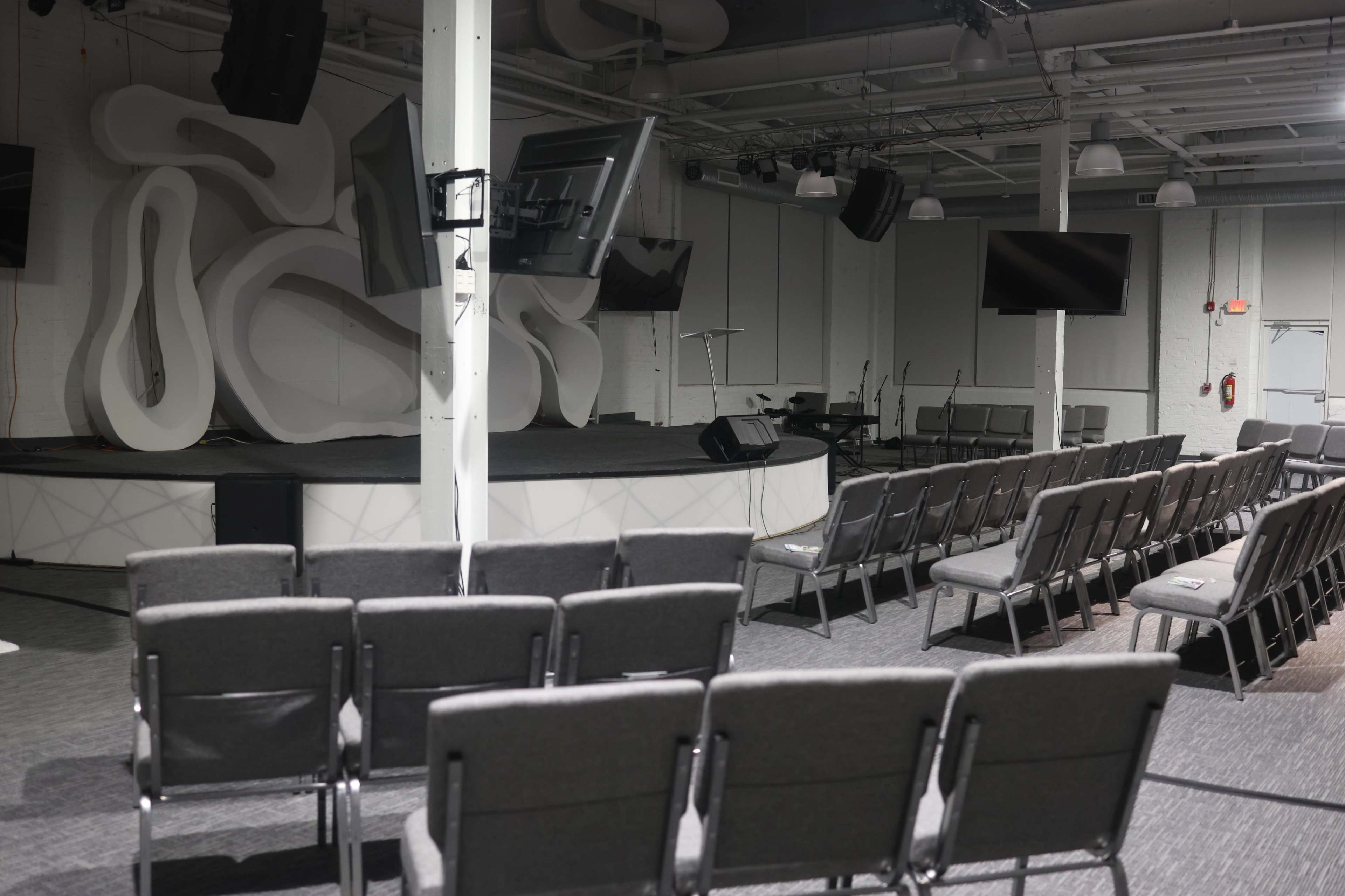 The image shows a modern indoor stage setup with a large abstract backdrop, a lectern, and rows of gray chairs arranged for an audience.