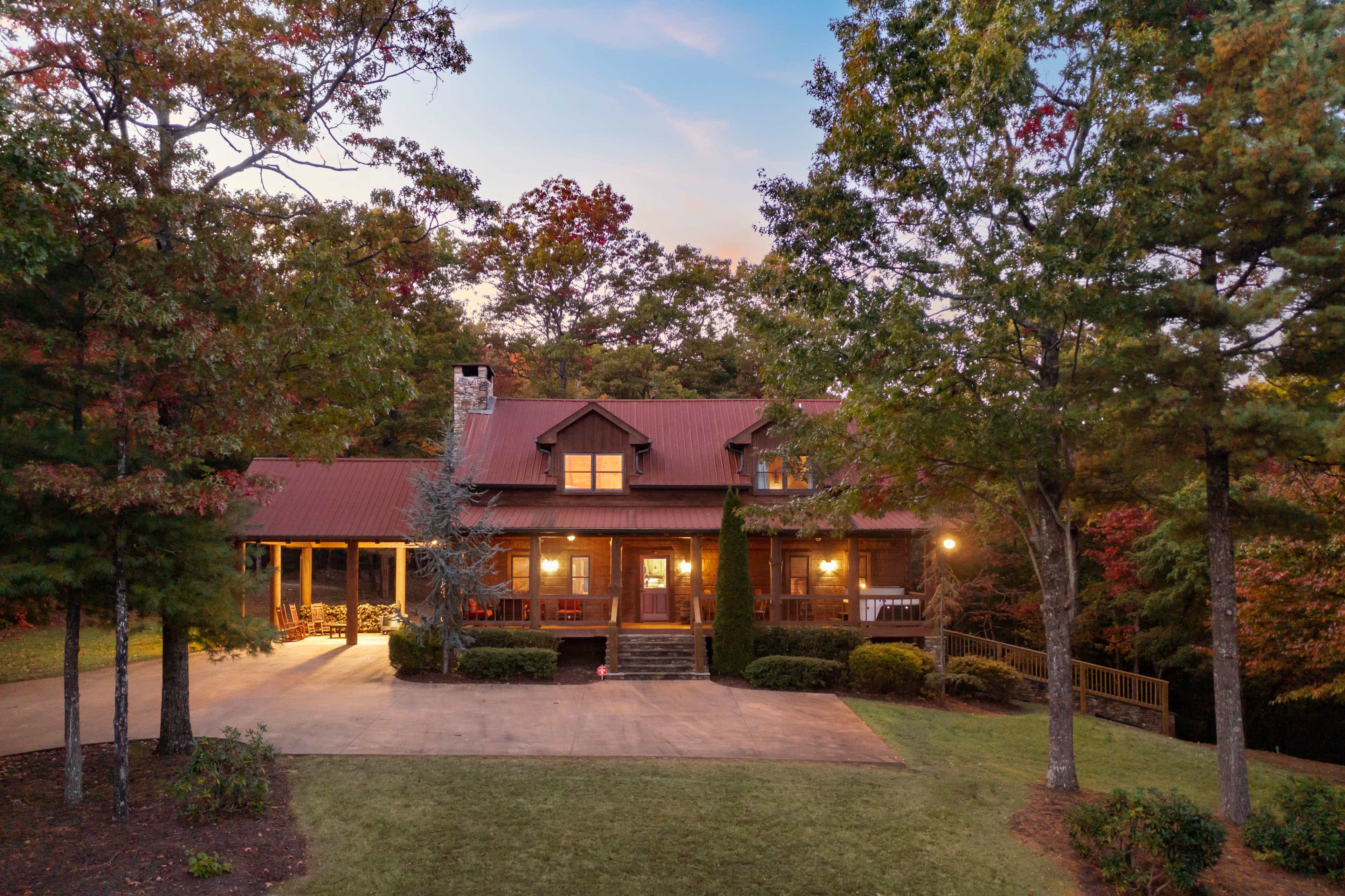 A large wooden house with a red metal roof is surrounded by trees and has a spacious front porch, situated on a well-manicured lawn.