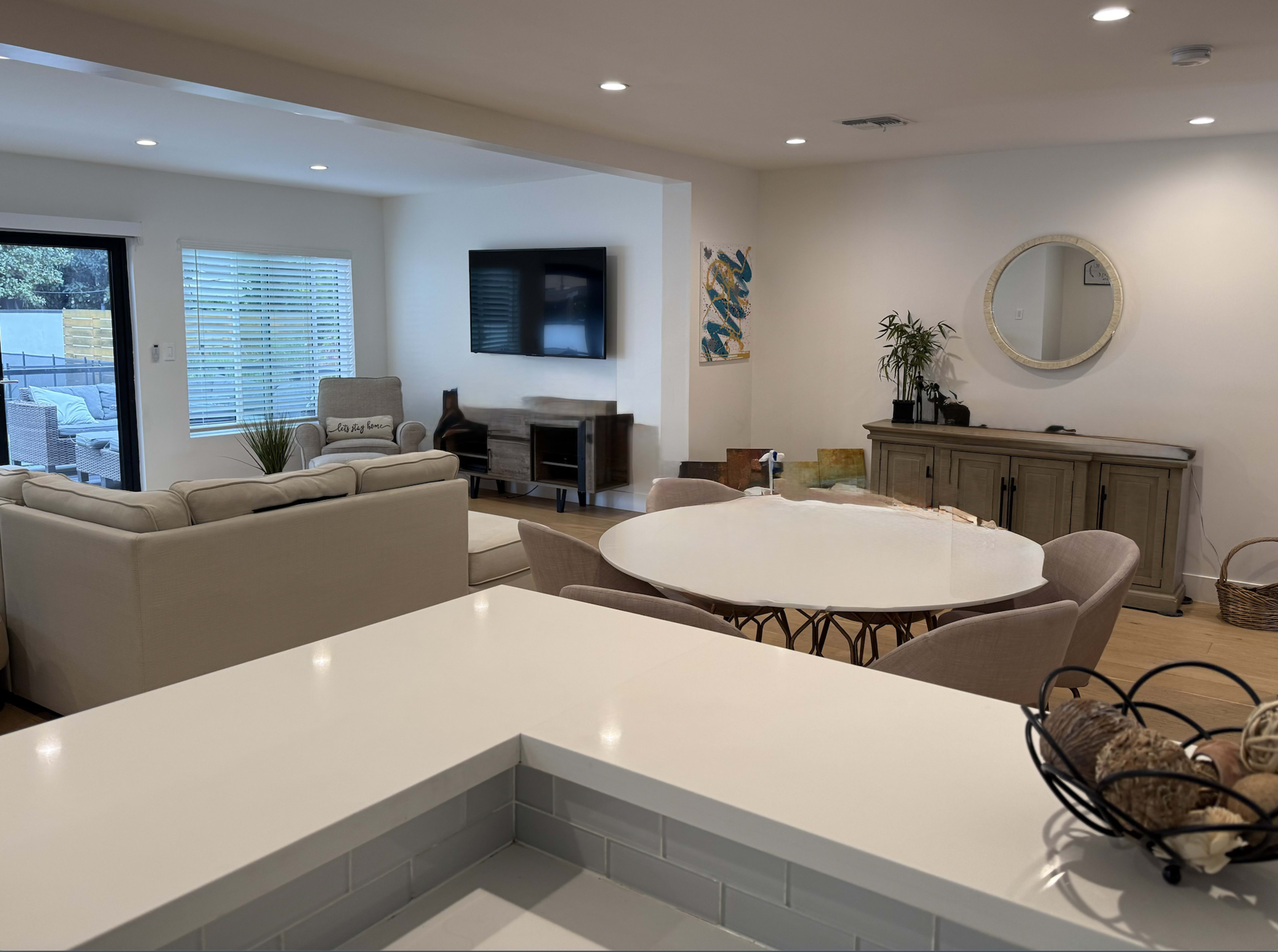 The image shows a modern living space featuring a kitchen island in the foreground, a circular dining table with chairs, a living area with a sofa, a television mounted on the wall, and decorative elements like plants and artwork.