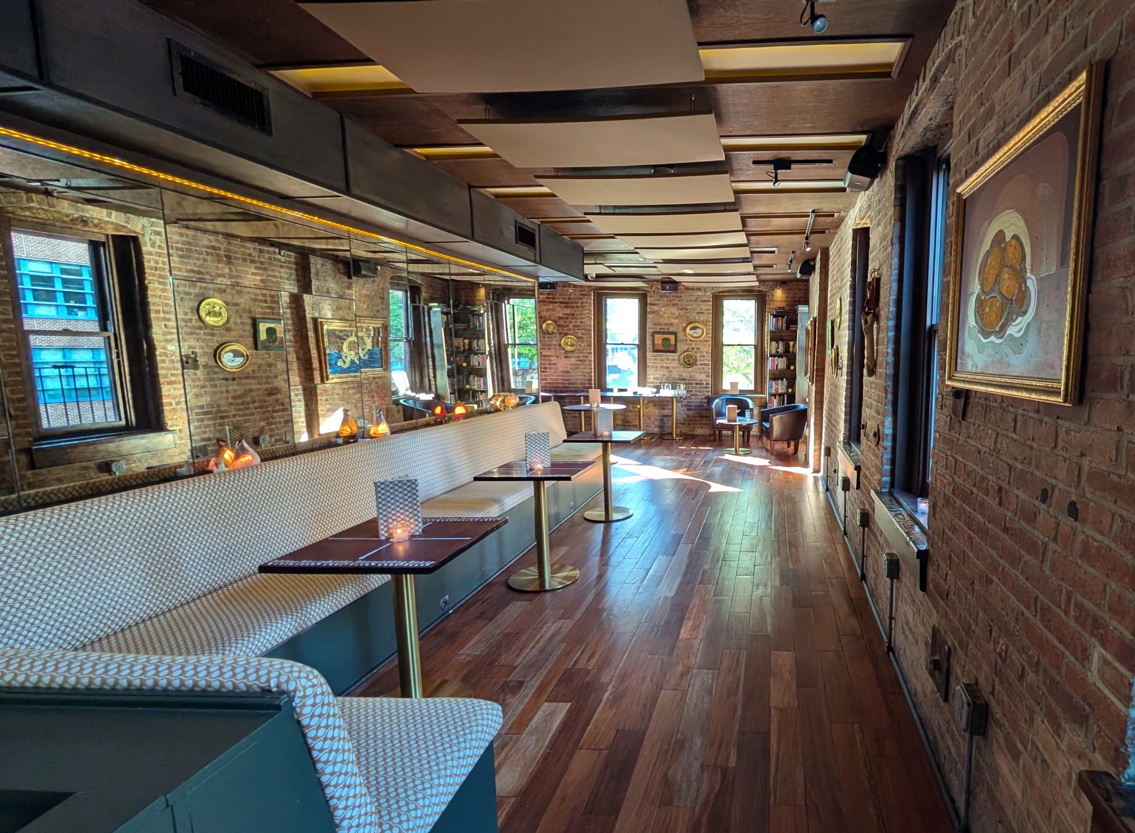 The image shows a spacious restaurant interior with exposed brick walls, wooden floors, and various seating arrangements, including a long banquette and tables set for dining.