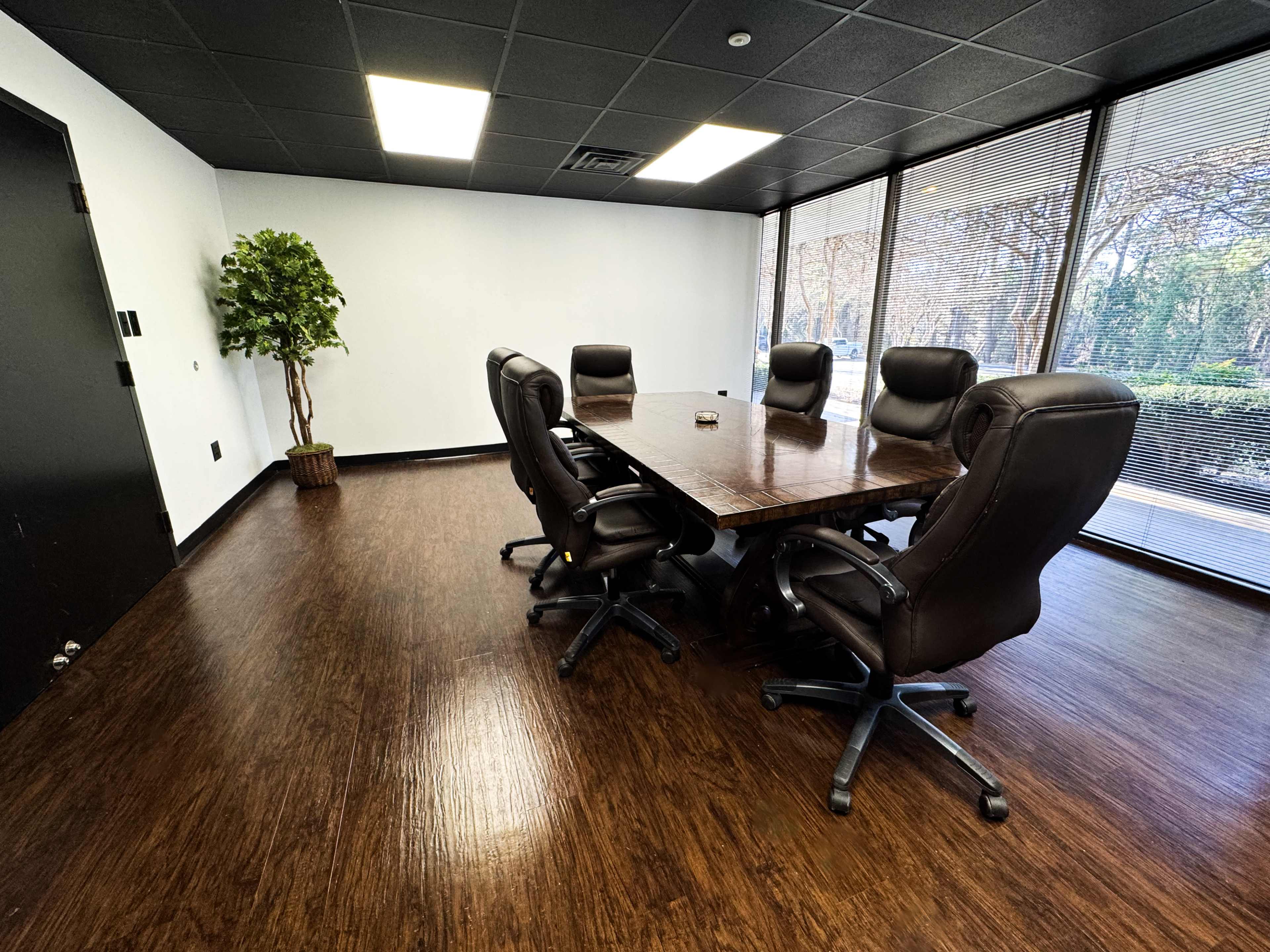 A conference room is furnished with a large wooden table surrounded by six black leather chairs, with a window providing a view of greenery outside.