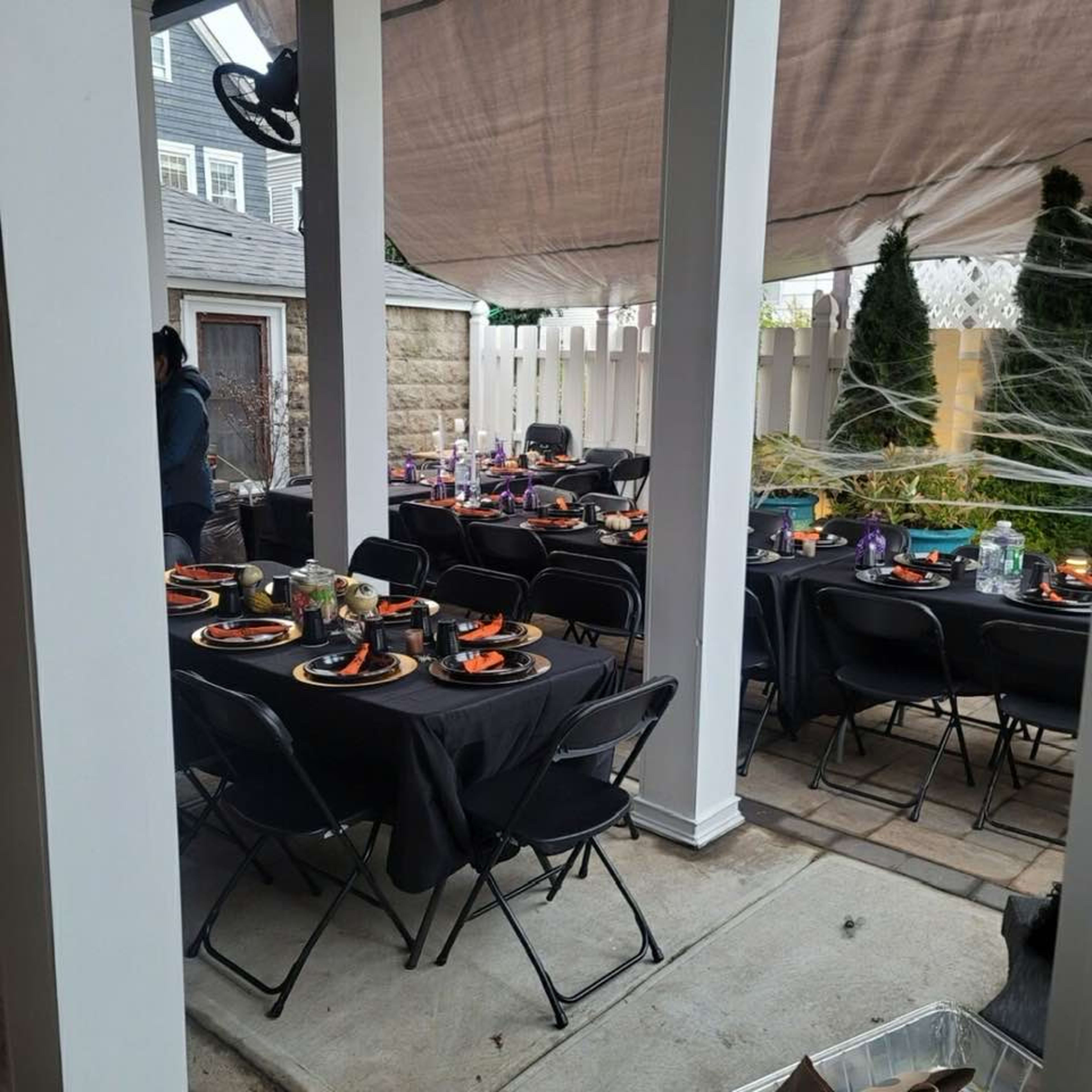 The image shows a covered outdoor area set up with multiple black tables and chairs, decorated with Halloween-themed tableware and surrounded by an enclosure.