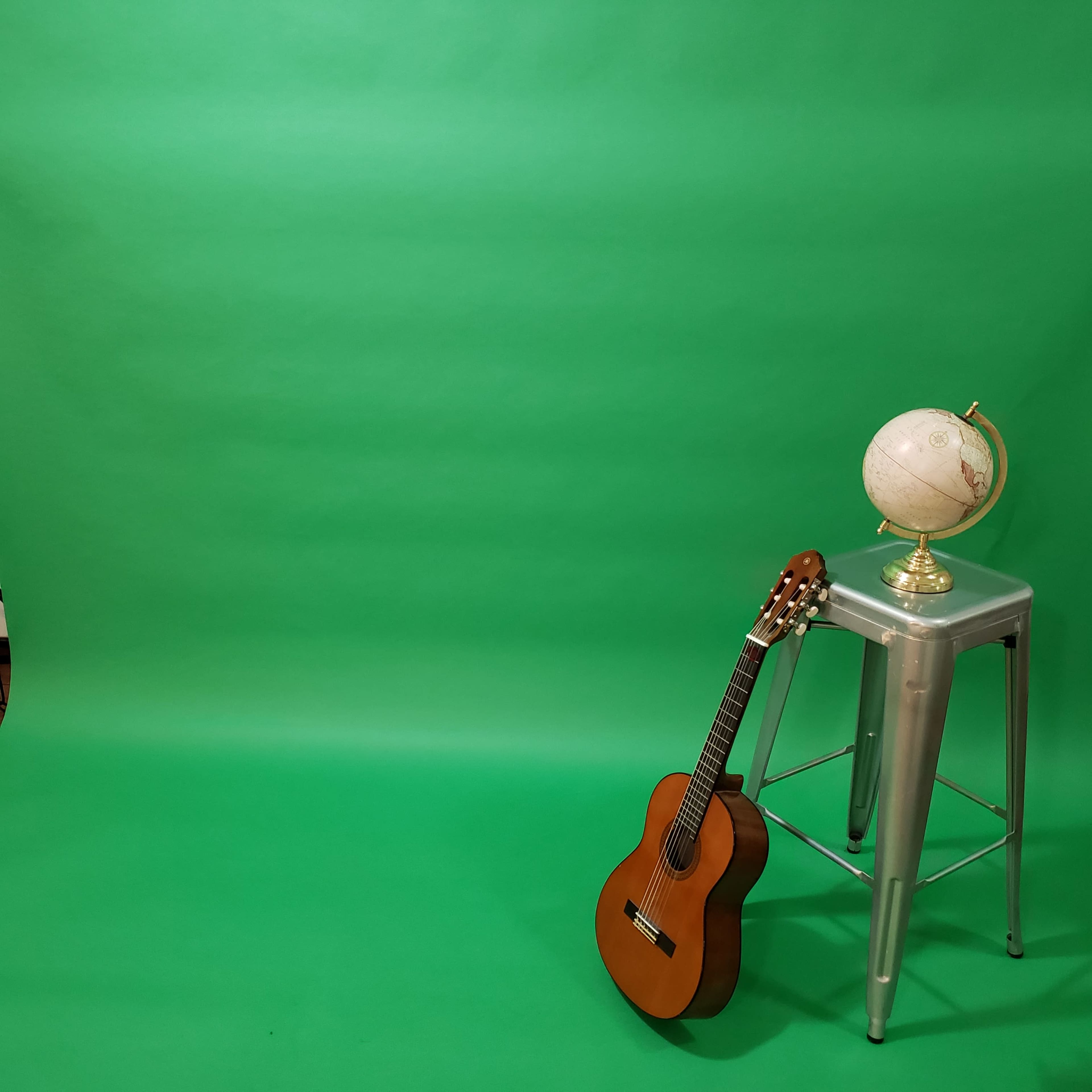 A wooden guitar rests against a metallic stool beside a globe on a green backdrop.