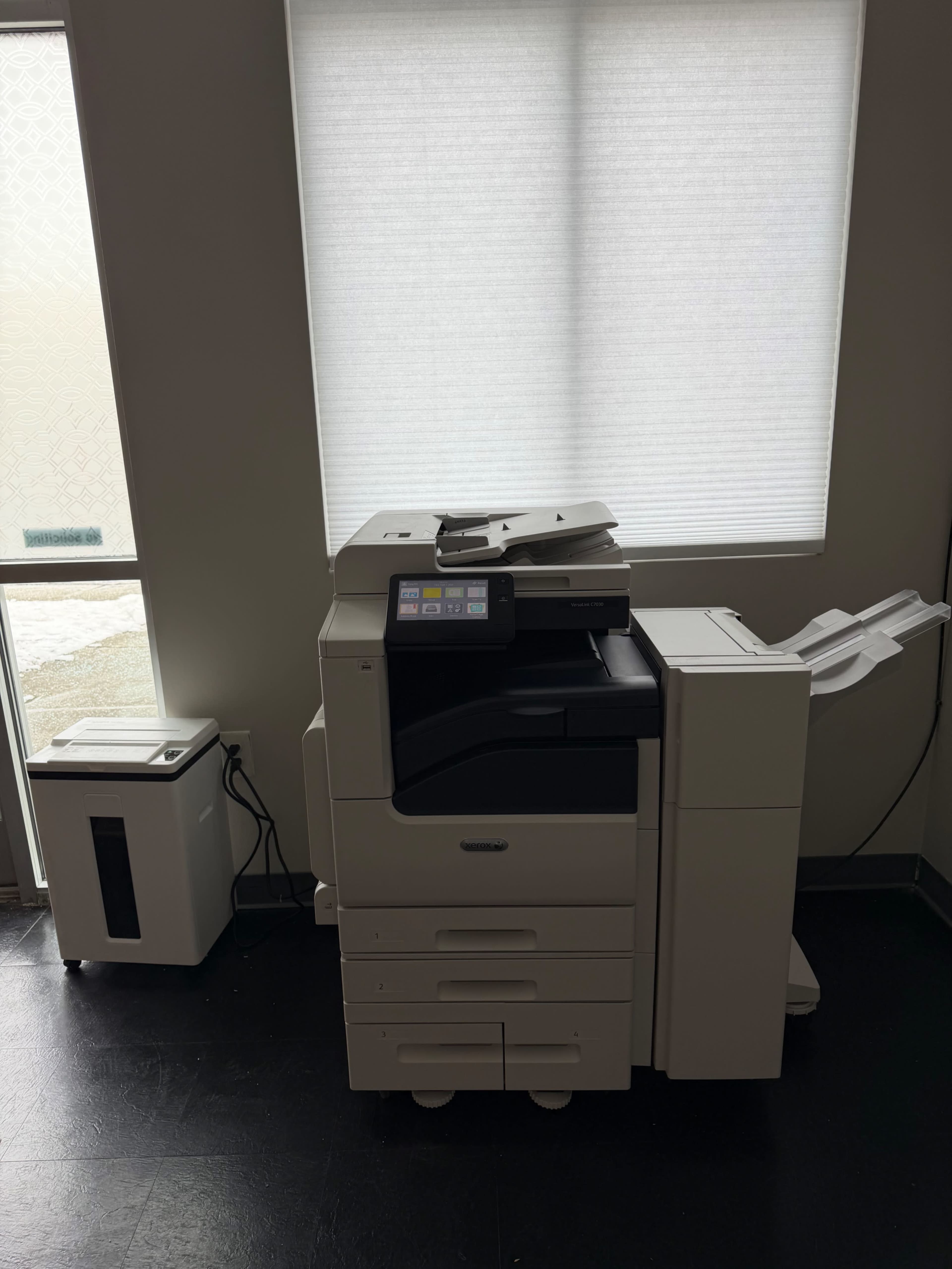 A large multifunction printer is positioned against a wall near a window, with a small paper shredder beside it.