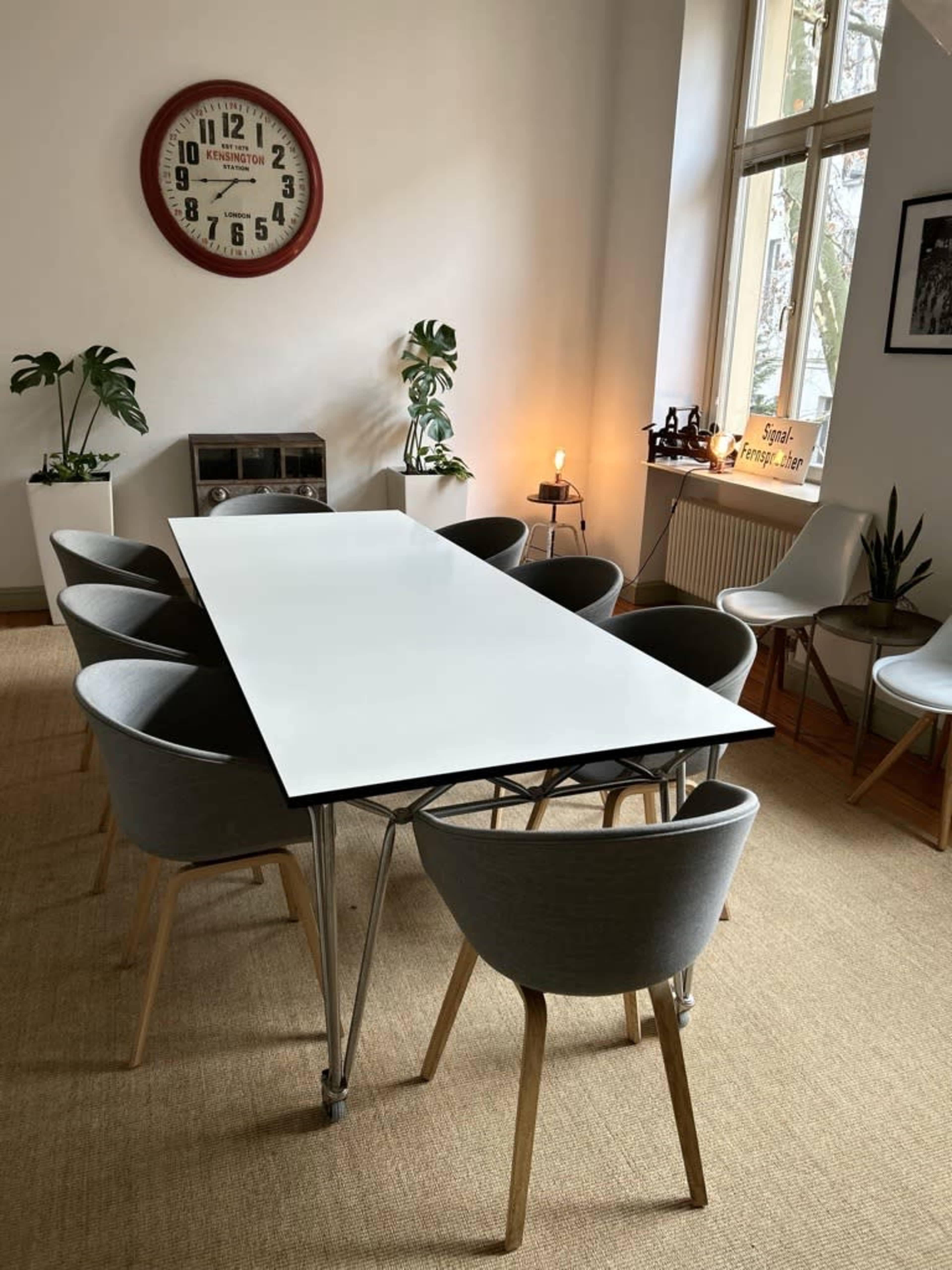 A long white table is surrounded by eight gray chairs in a well-lit meeting room.