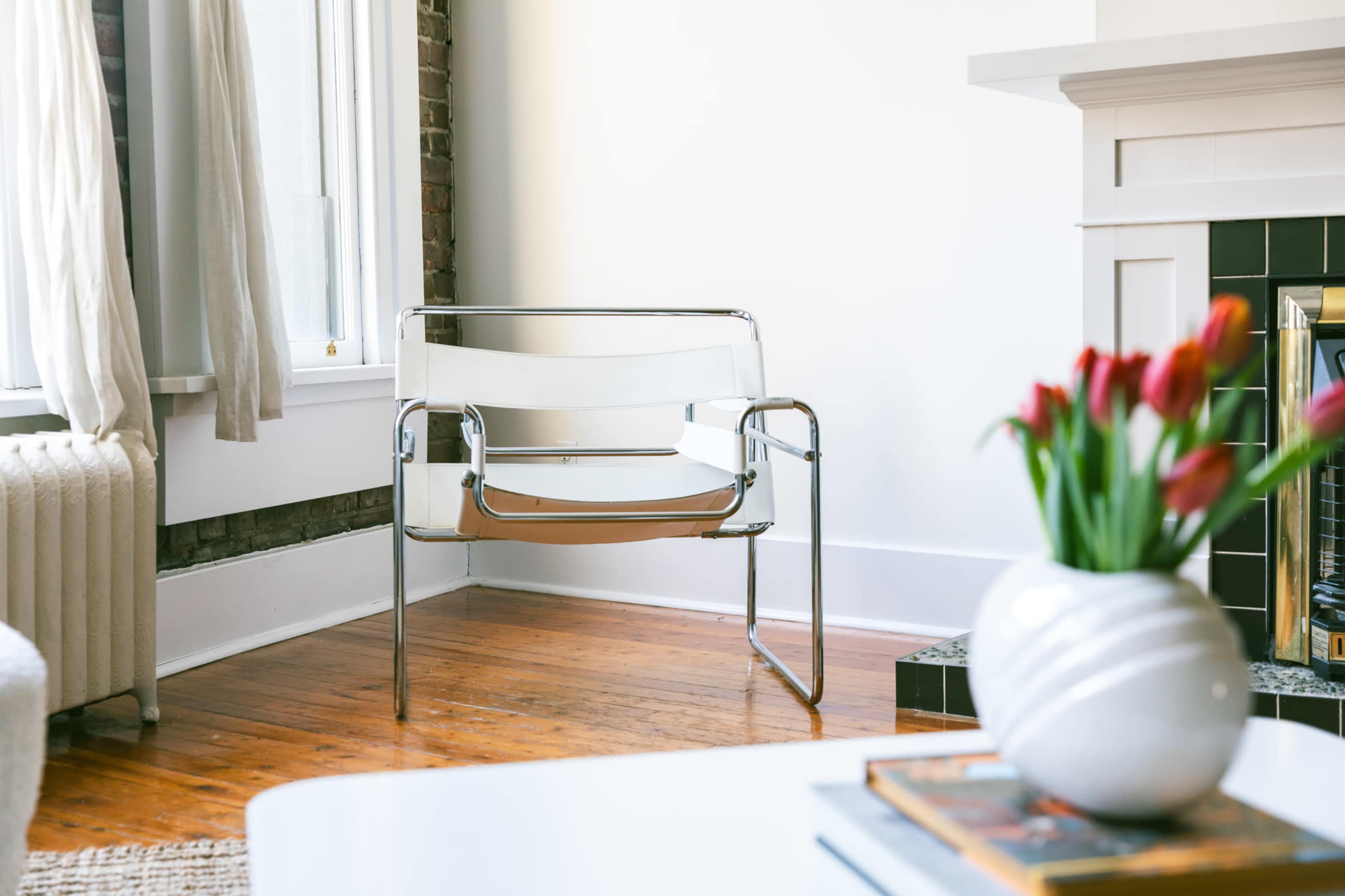 A modern chrome chair with a clear seat and backrest is positioned in a corner of a room with hardwood flooring and a white wall.