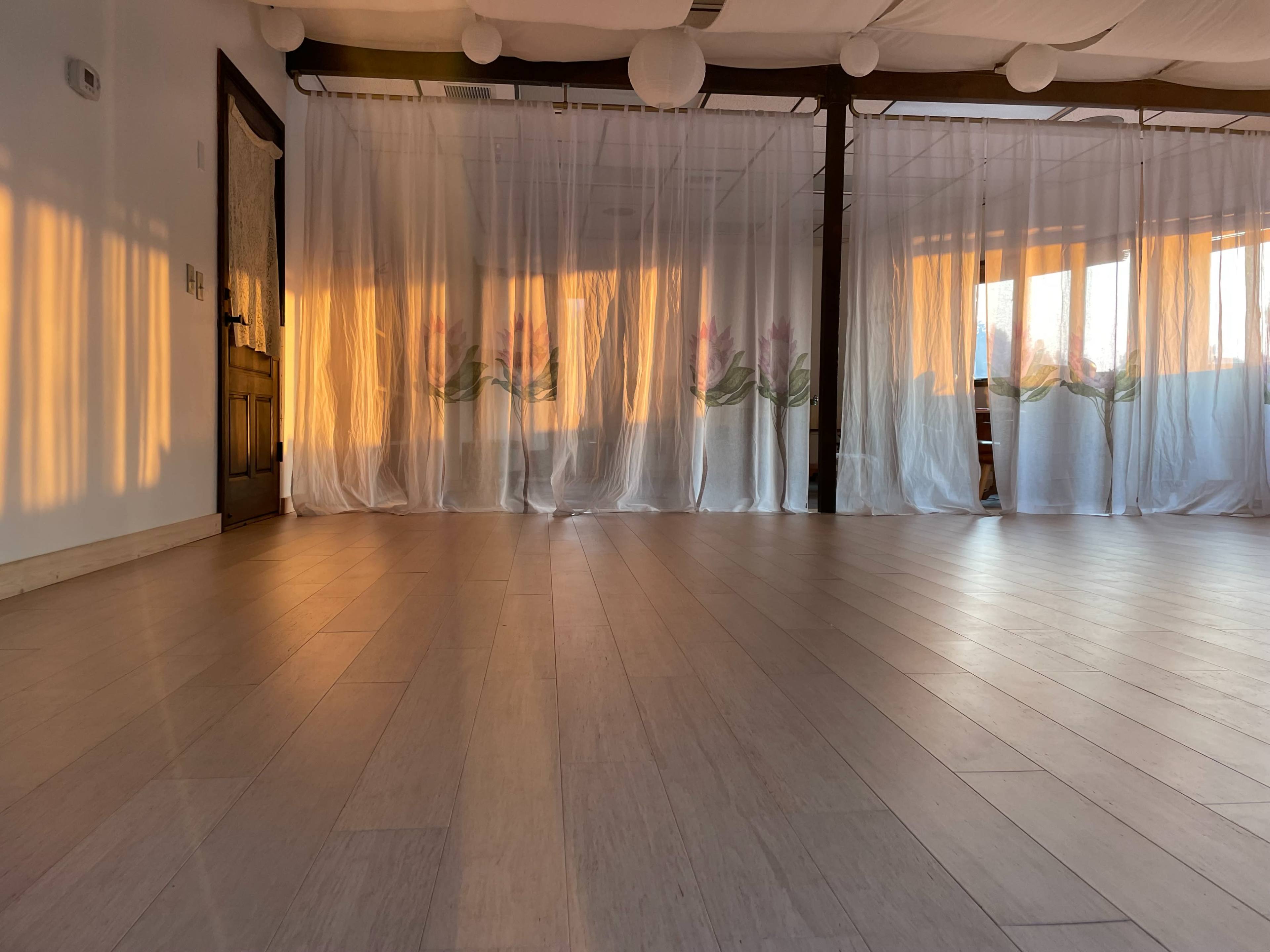 A spacious room with wooden flooring, sheer white curtains adorned with floral designs, and warm sunlight streaming through them.