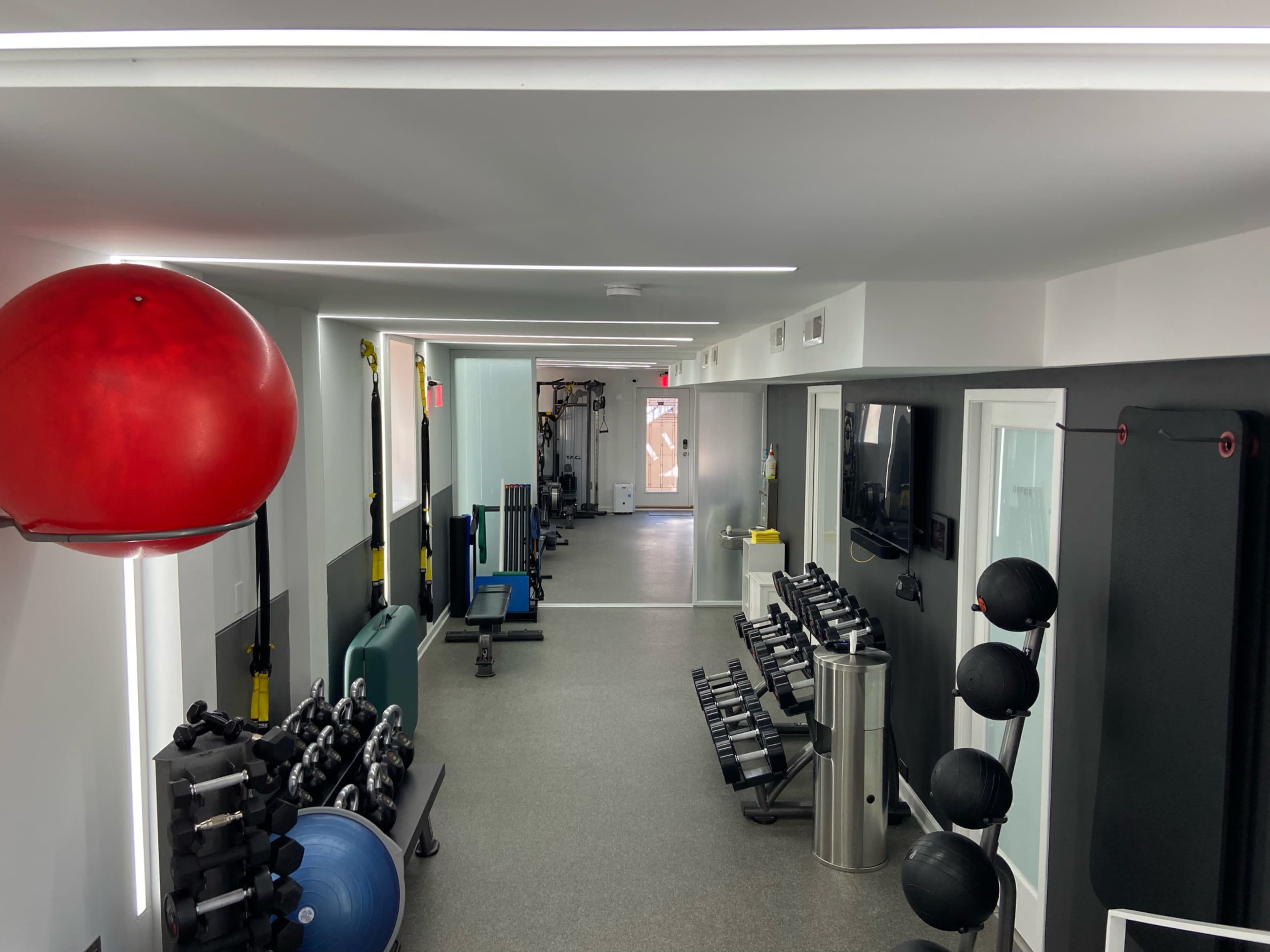 A modern gym interior featuring various fitness equipment, including dumbbells, a stability ball, and an exercise bench, arranged along a long hallway.