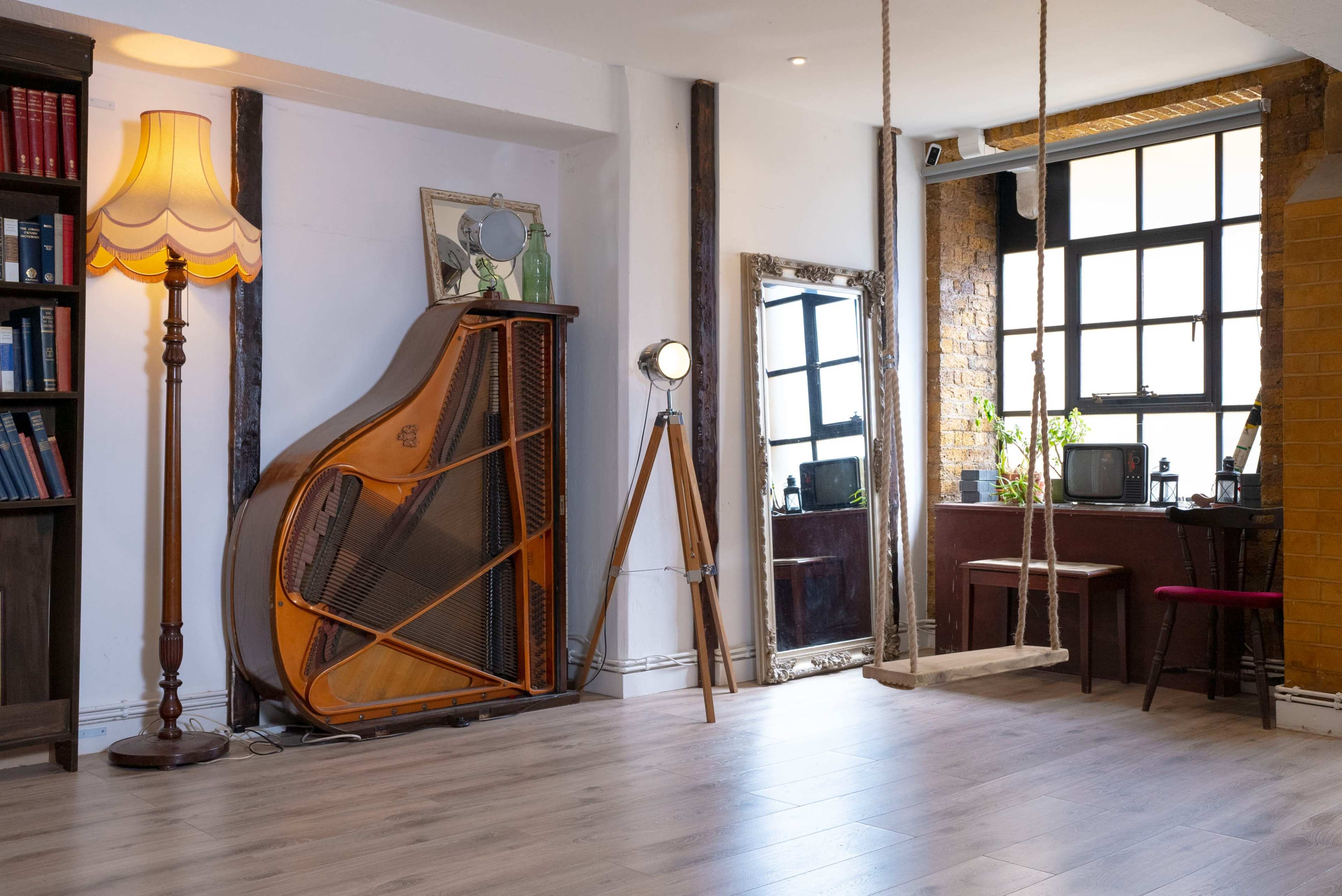 A spacious, well-lit interior features a grand piano against the wall, a floor lamp, a mirror, and a swing, with bookshelves and vintage decor.