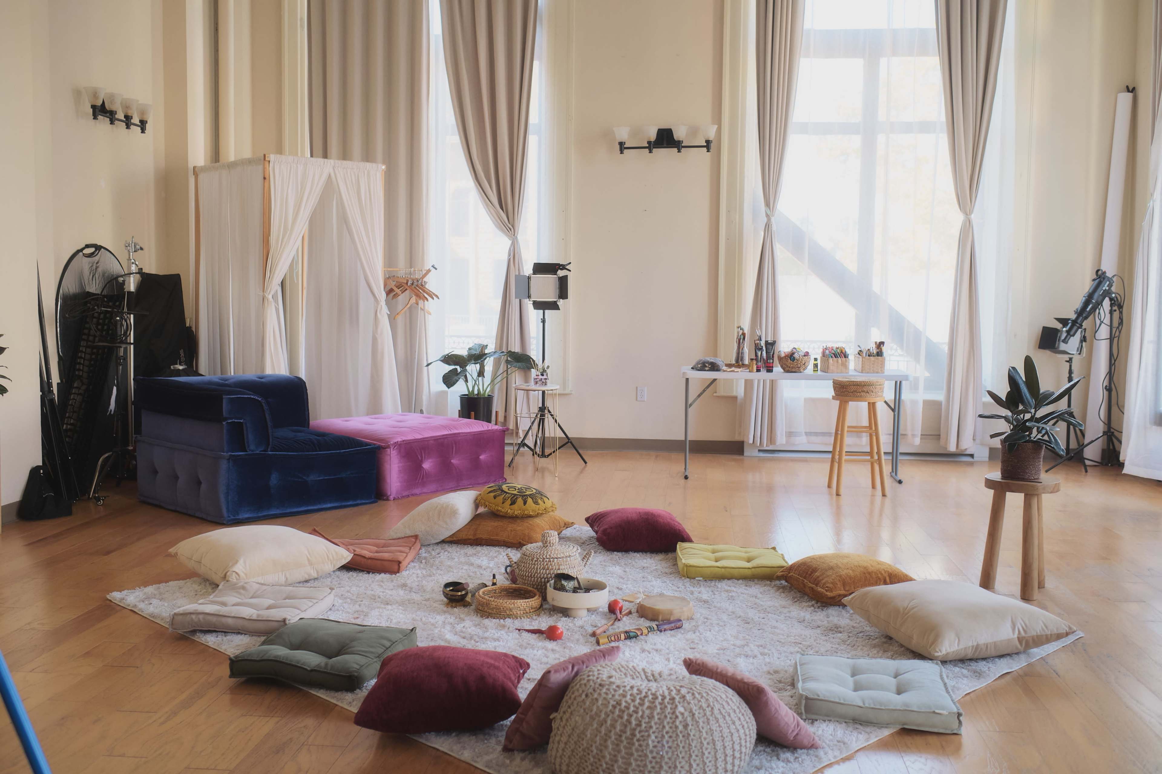 The image shows a spacious interior with a circular arrangement of colorful cushions and a soft rug, surrounded by large windows and various decorative items.