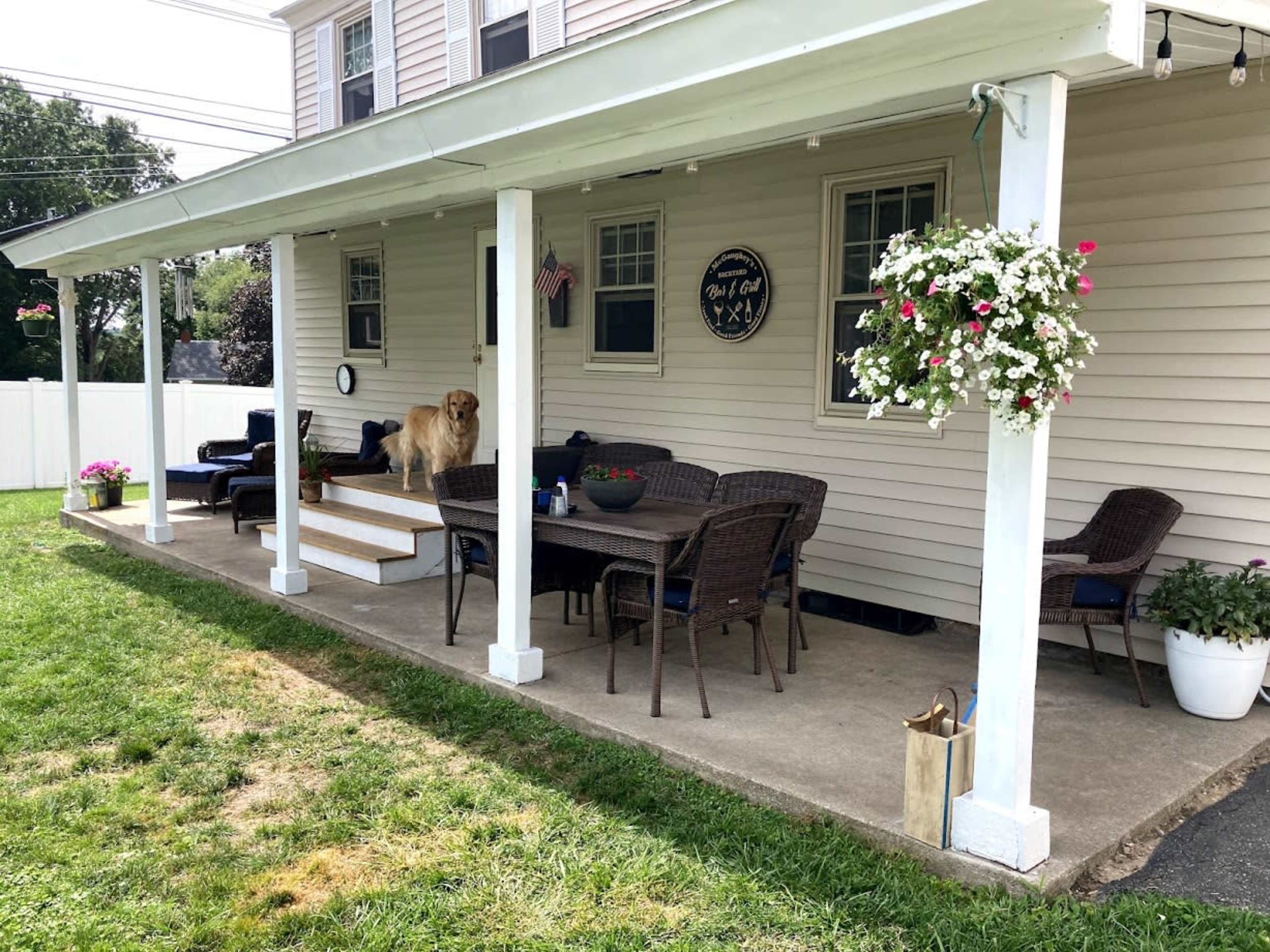 A covered patio with a dining table, chairs, a golden retriever, and hanging flower pots.