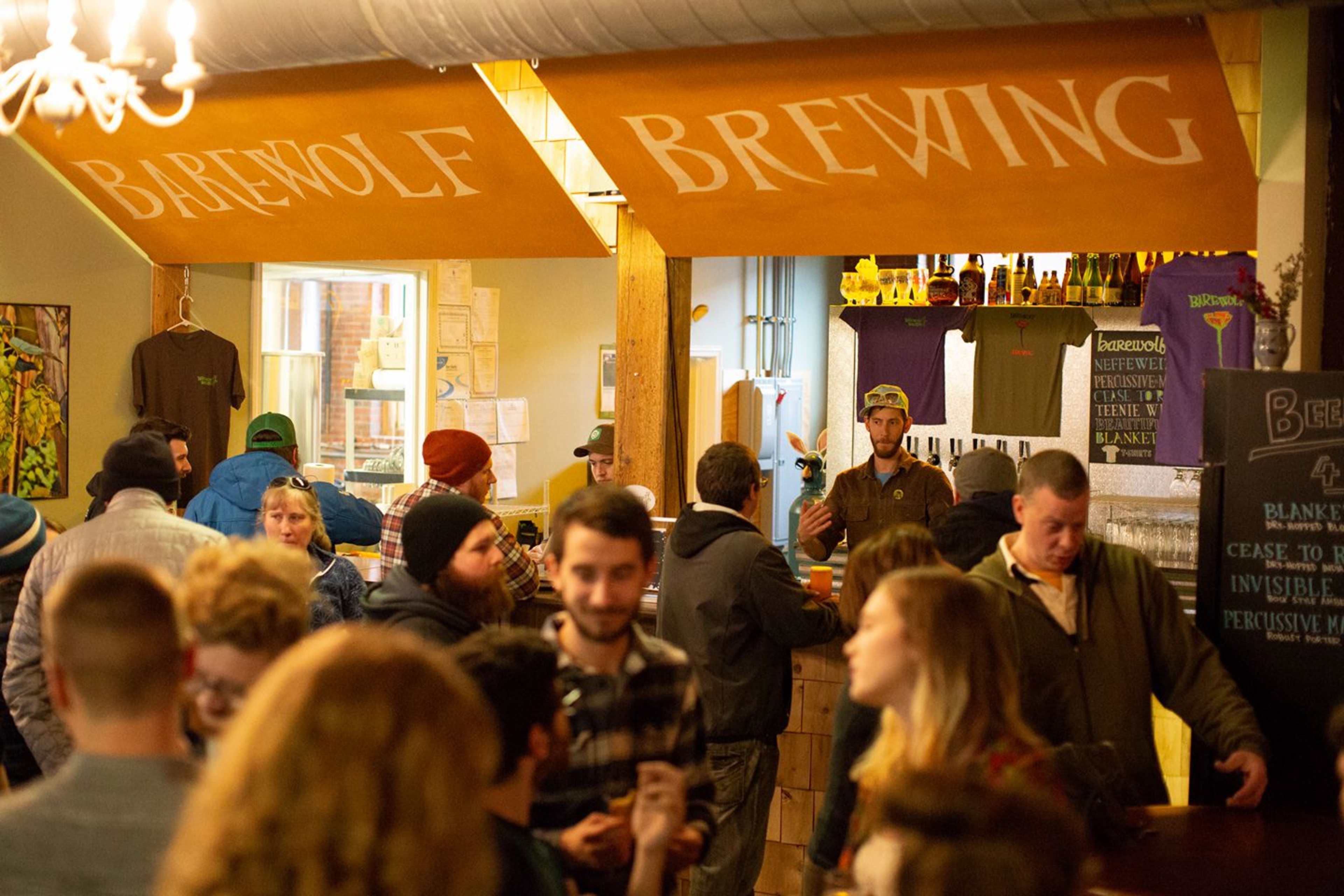 A crowded brewery taproom with patrons mingling at the bar and merchandise displayed on the walls.