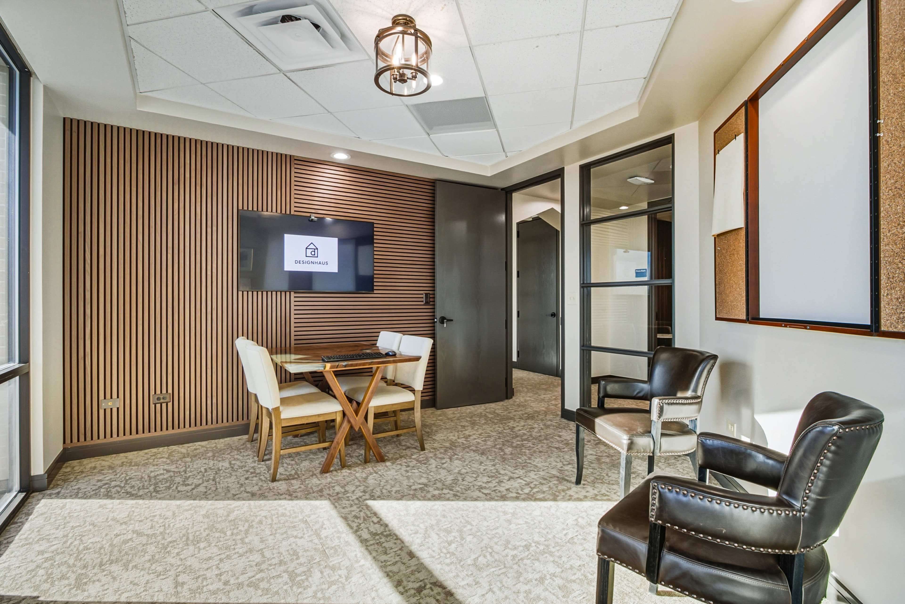 The image shows a small, modern office meeting room with a wooden accent wall, a table with four chairs, and a television mounted on the wall.