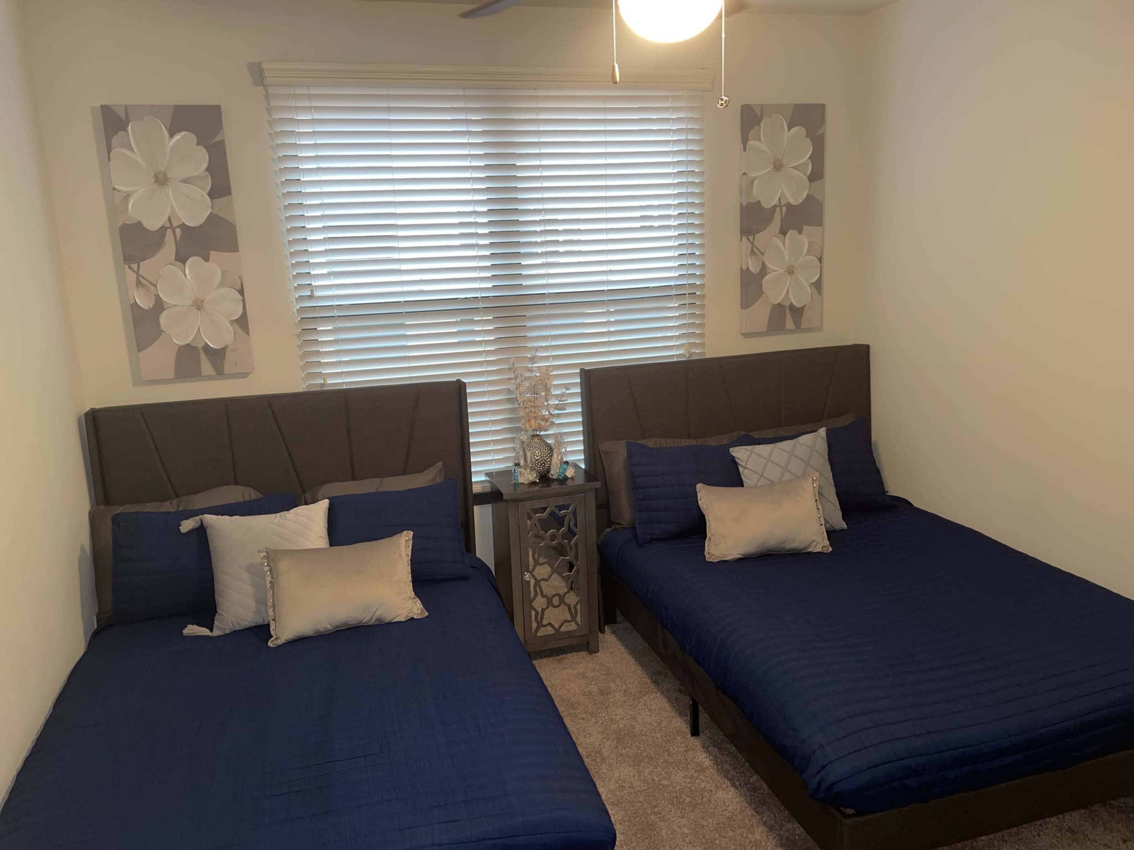 A bedroom with two twin beds covered in blue quilts, framed by light-colored walls and decorated with flower artwork above each bed.
