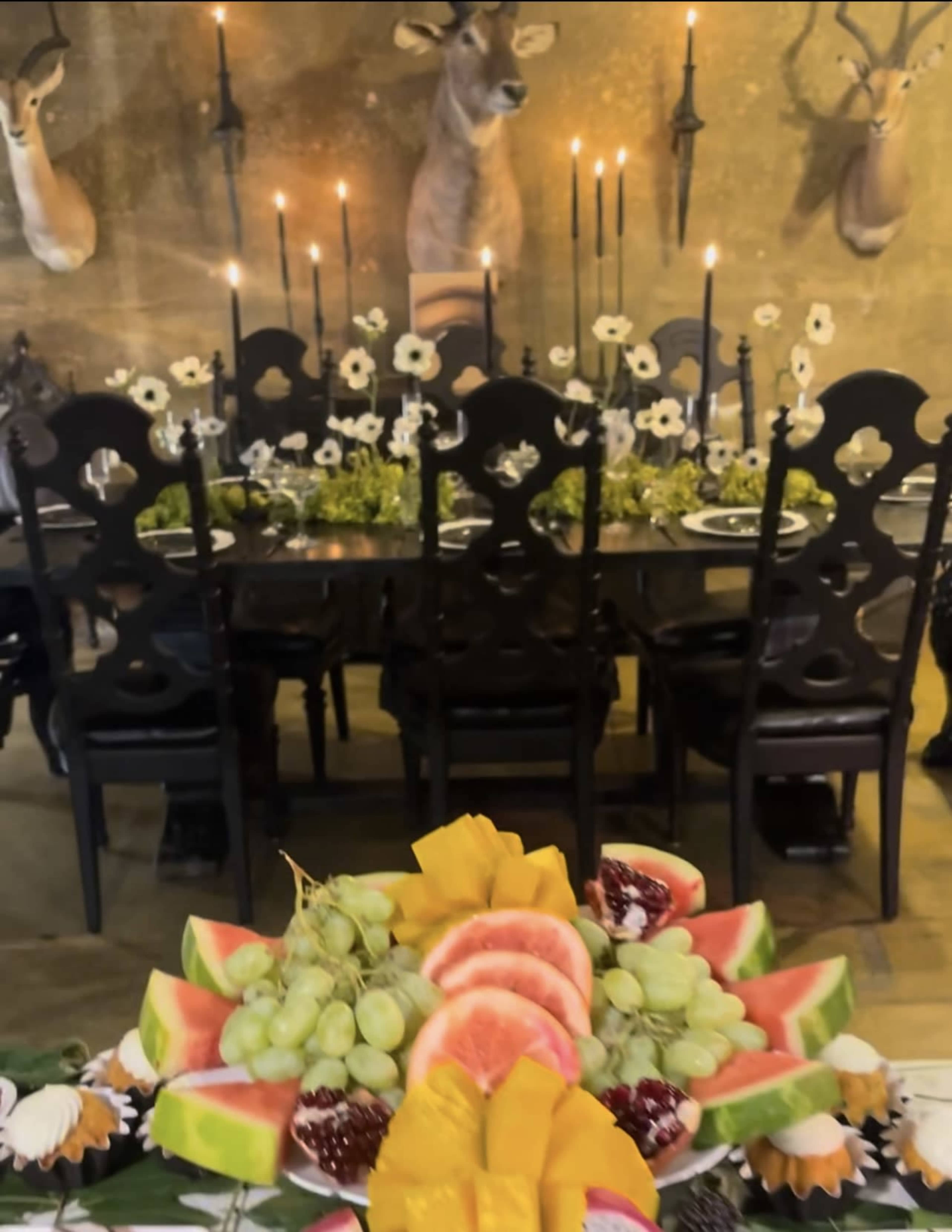The image shows a dining table set with black chairs, adorned with candles and flowers, while a colorful fruit platter in the foreground features various sliced fruits.