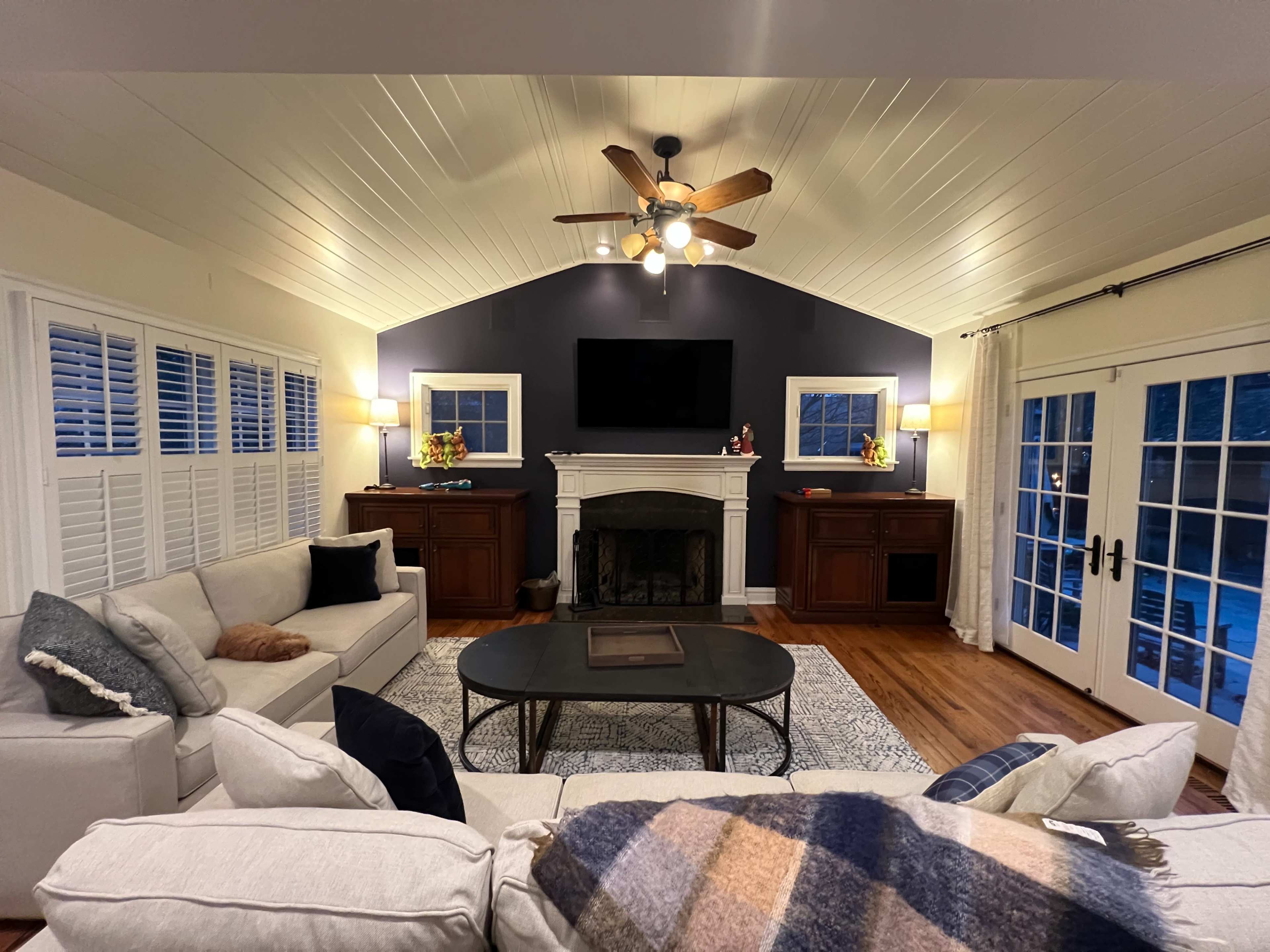 A spacious living room features a vaulted ceiling, a dark accent wall with a mounted TV, and light-colored furniture arranged around a coffee table.