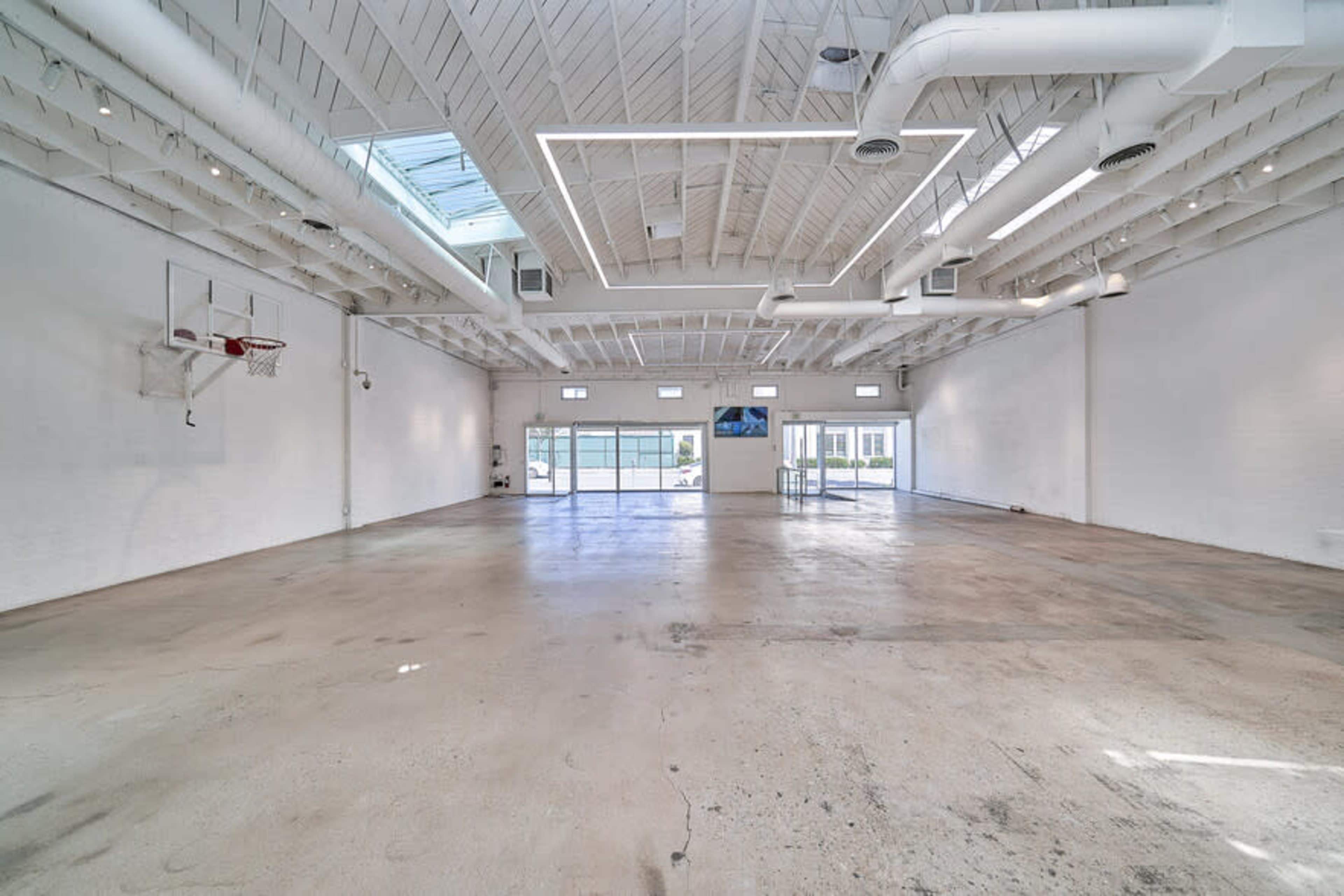A spacious, empty indoor area features a basketball hoop on one wall and large windows letting in natural light.