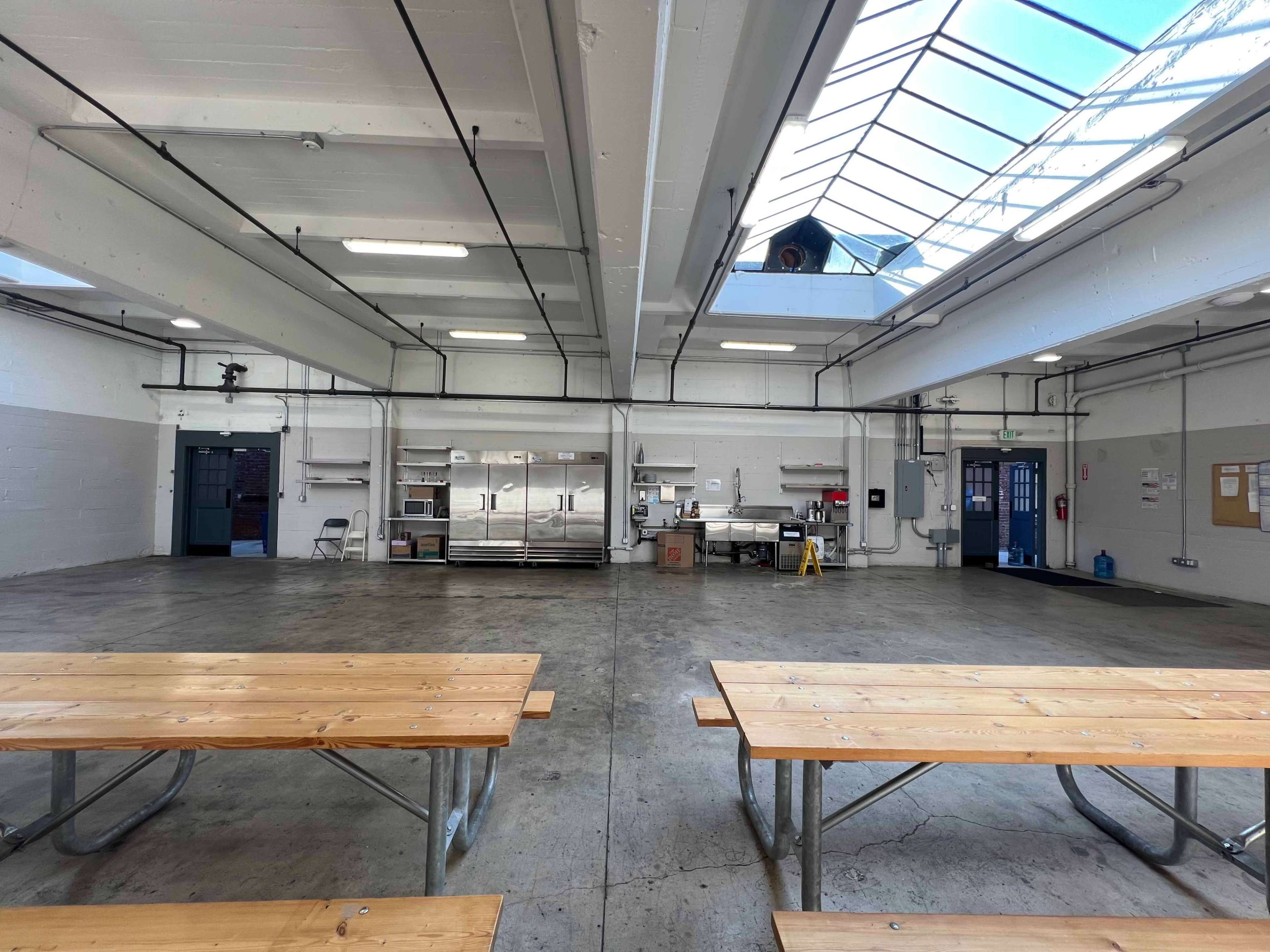 A spacious, industrial-style kitchen area with long wooden tables and stainless steel appliances, illuminated by natural light from skylights above.