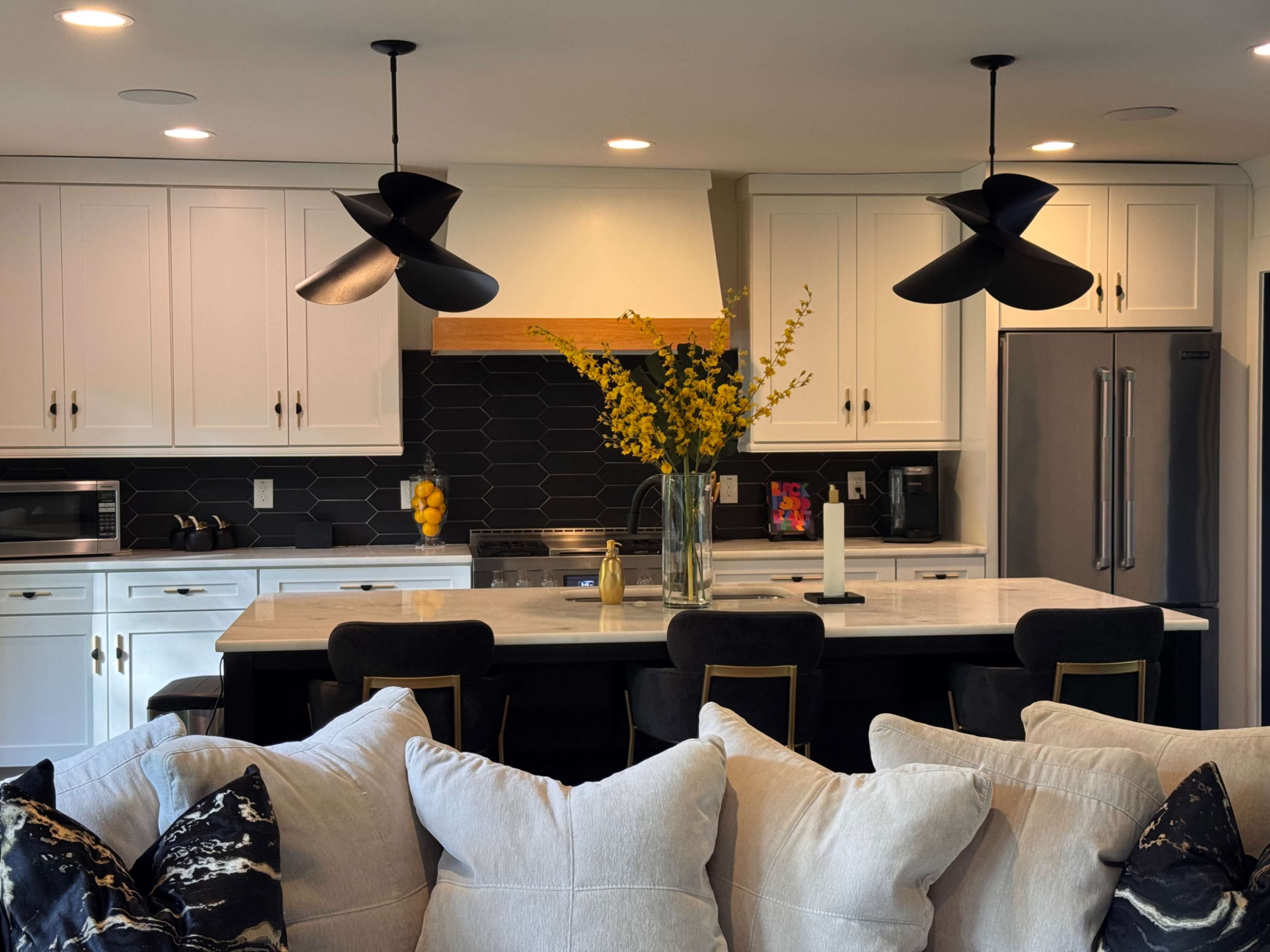 A modern kitchen features a central island with seating, black pendant lights, and a vase of yellow flowers atop a marble counter.