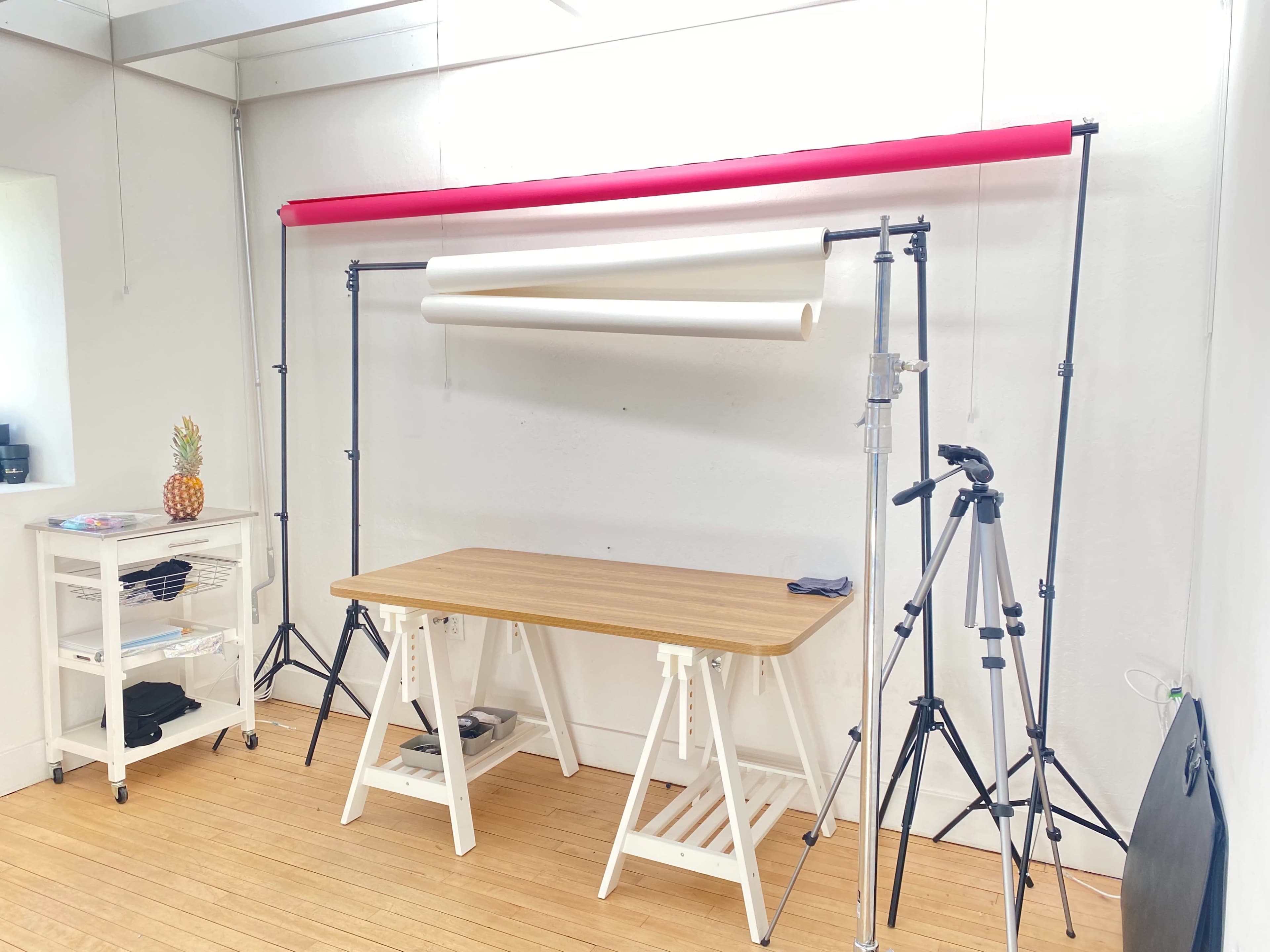 A photography studio setup features a wooden table, two tripods, and a backdrop support with red and white paper rolls.