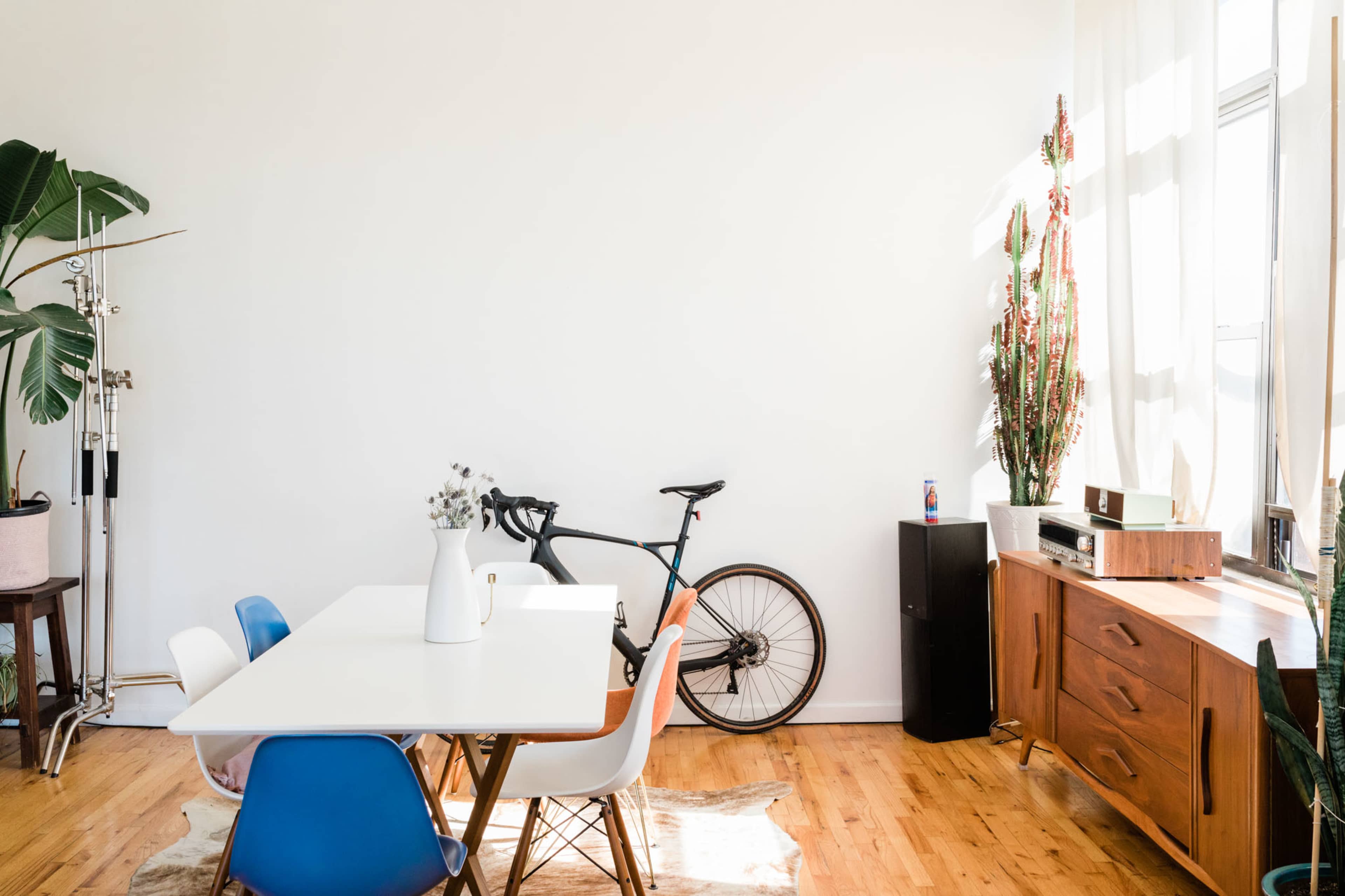 The image shows a bright room with a dining table, colorful chairs, a bicycle leaning against the wall, and a wooden sideboard.