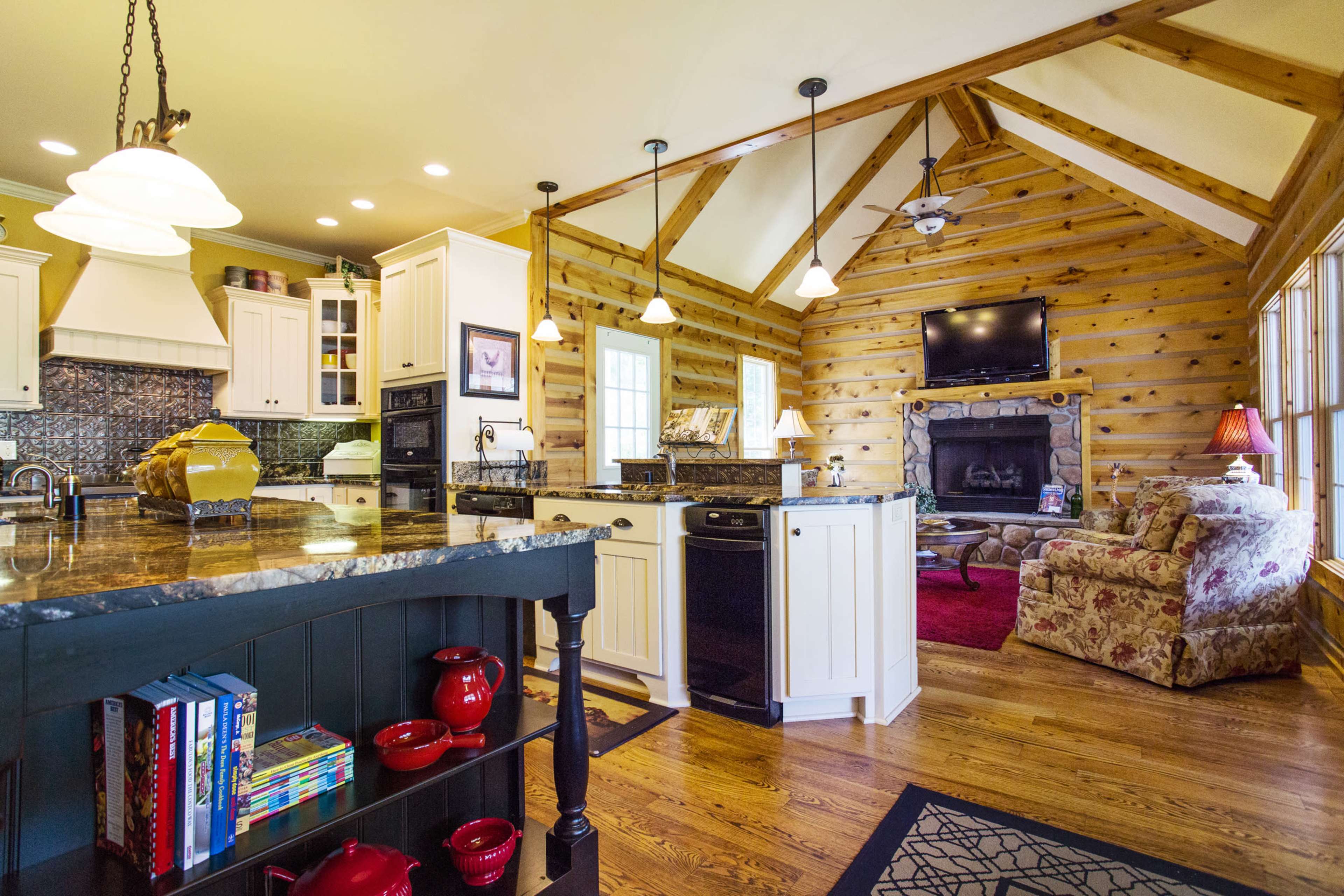 A rustic kitchen with wooden walls, an island with a dark granite countertop, and a cozy living area featuring a stone fireplace and a floral-patterned chair.