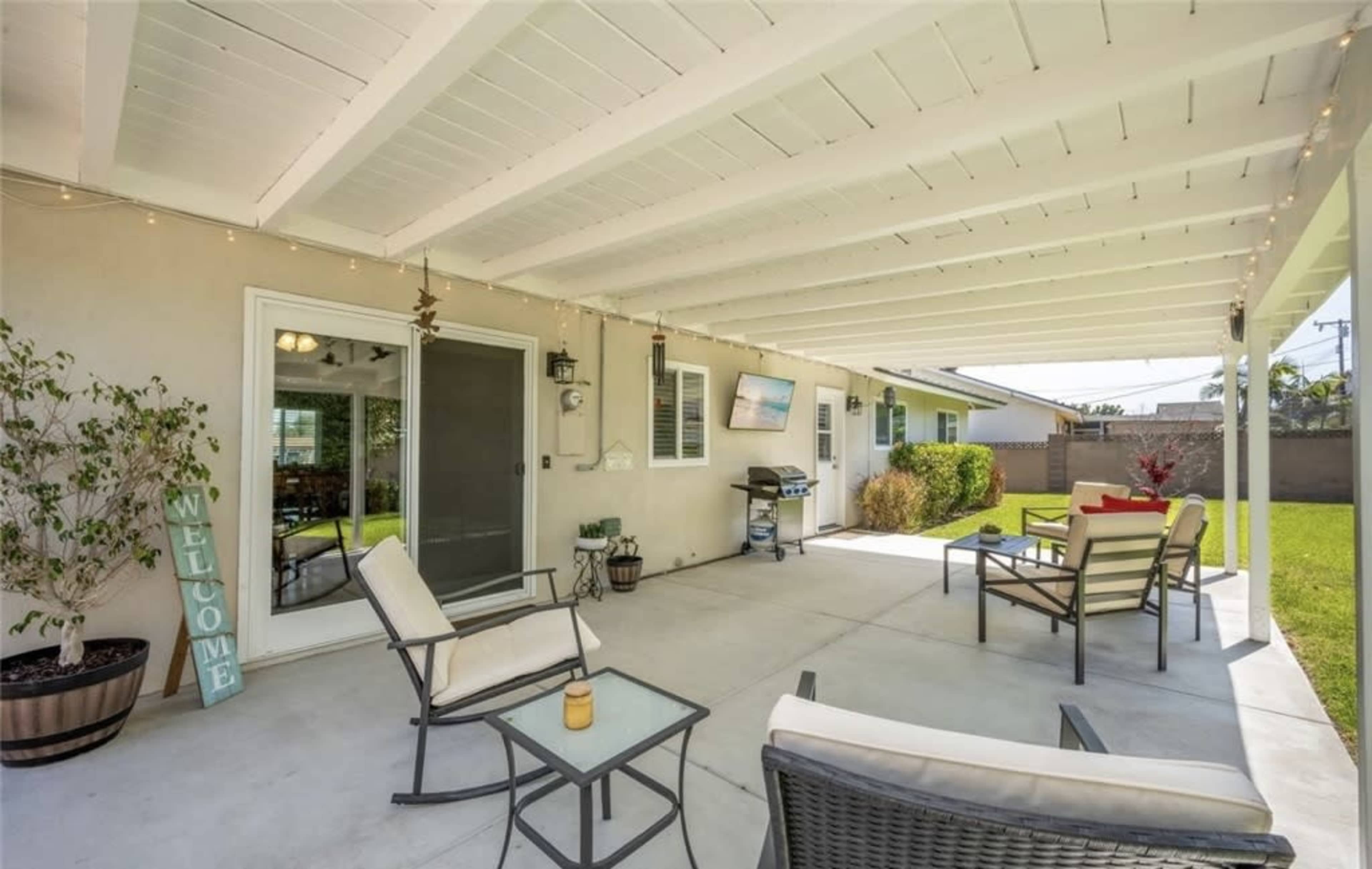 The image shows a covered patio area with several chairs, a small table, and a barbecue grill, surrounded by a lawn and greenery.