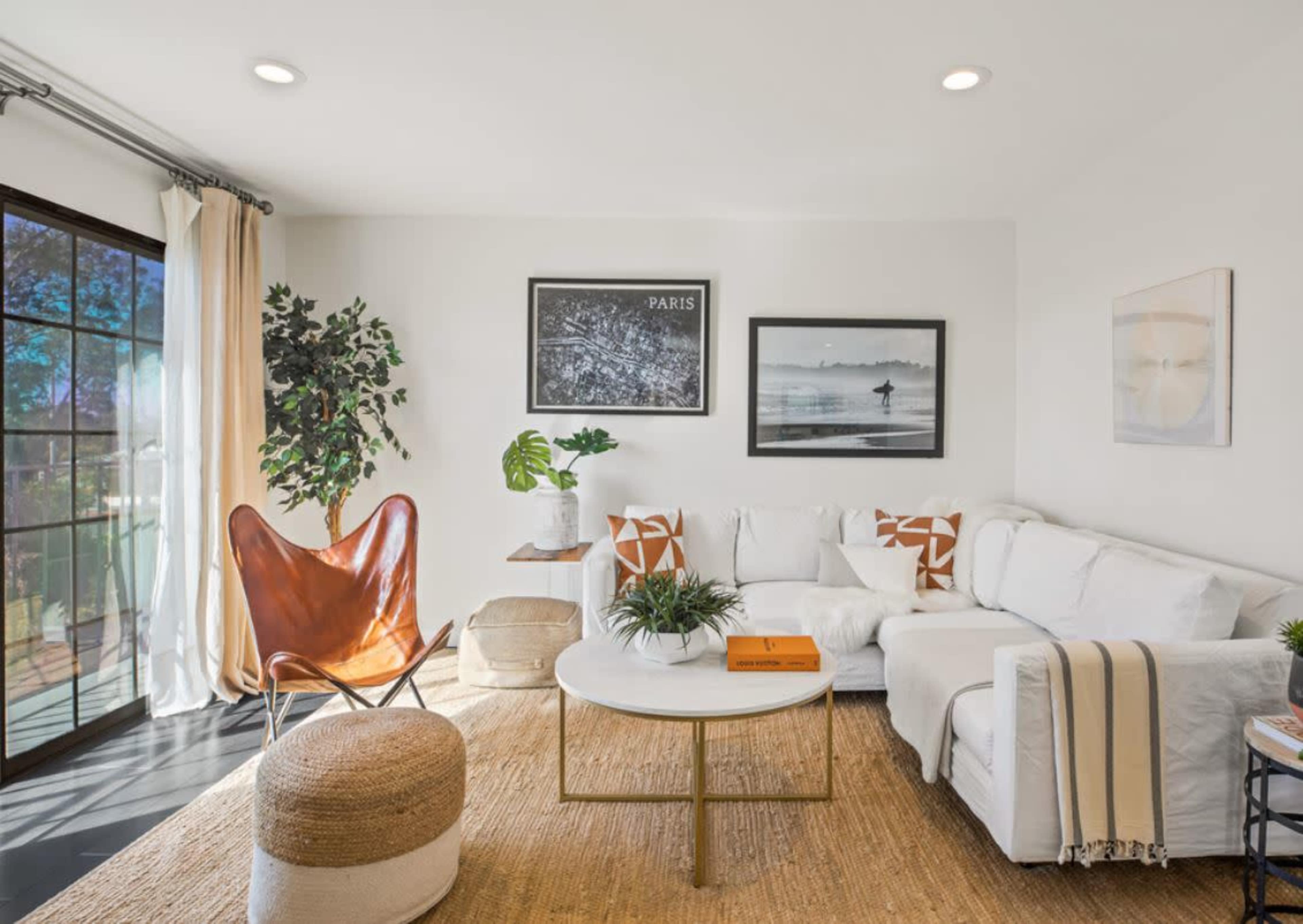 The living room features a white sectional sofa, a round coffee table, a butterfly chair, and decorative plants, all arranged around a large window with natural light.