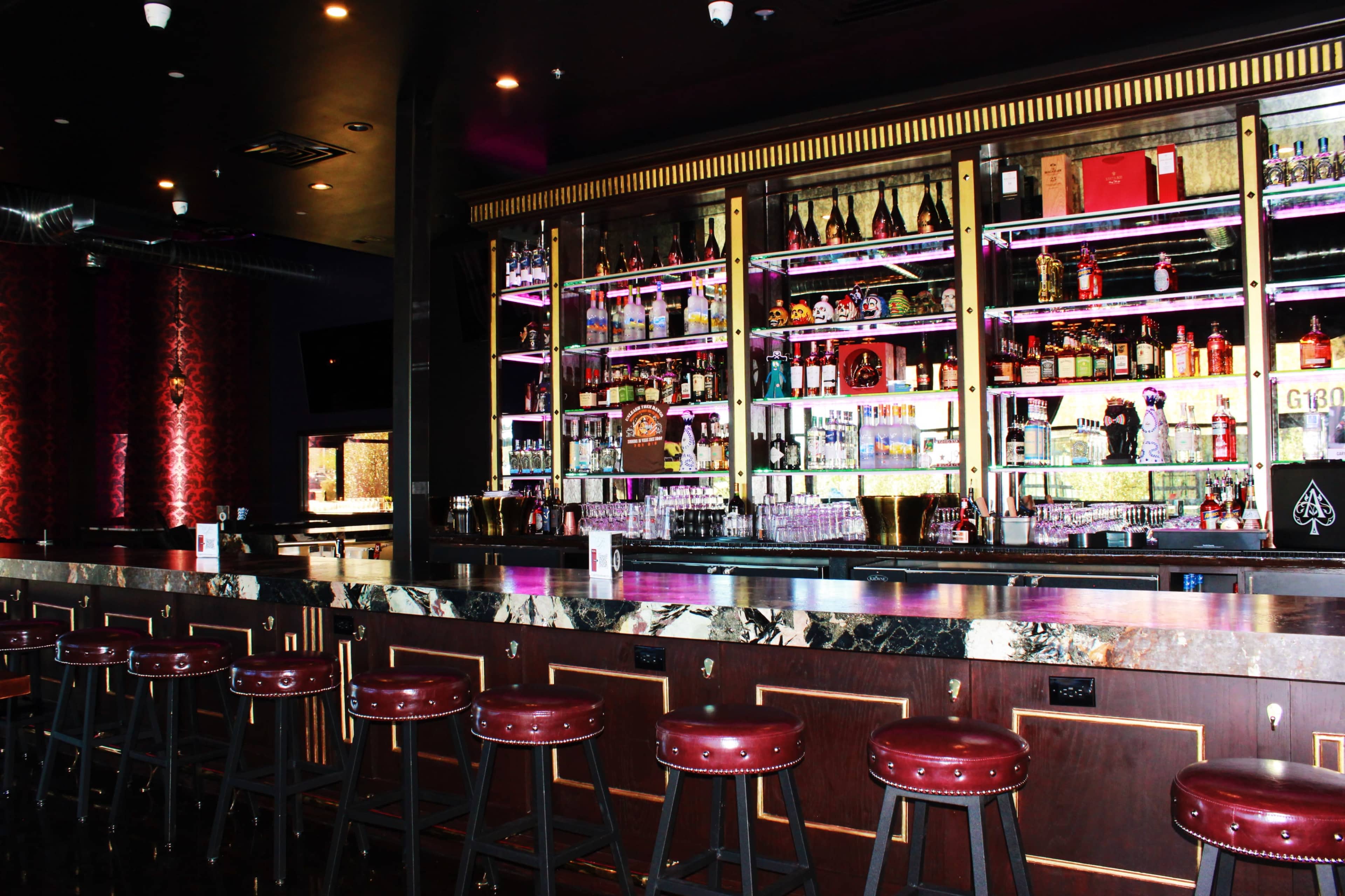 The image shows a well-lit bar area with a polished marble counter and high stools, featuring shelves of various liquor bottles illuminated by purple backlighting.
