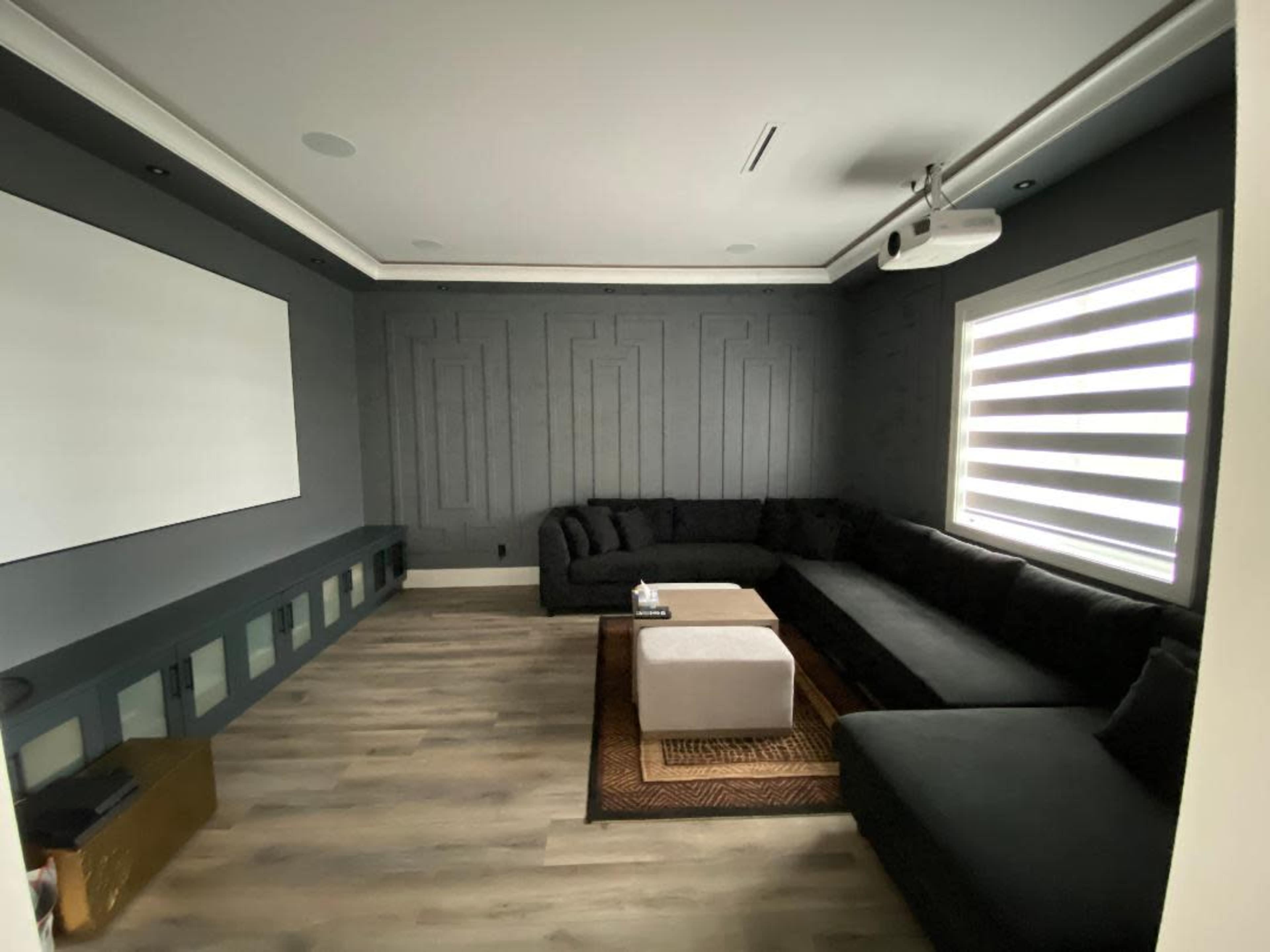 A modern living room features a large dark sectional sofa, a white coffee table, and a projector screen mounted on the wall, surrounded by minimalist decor.
