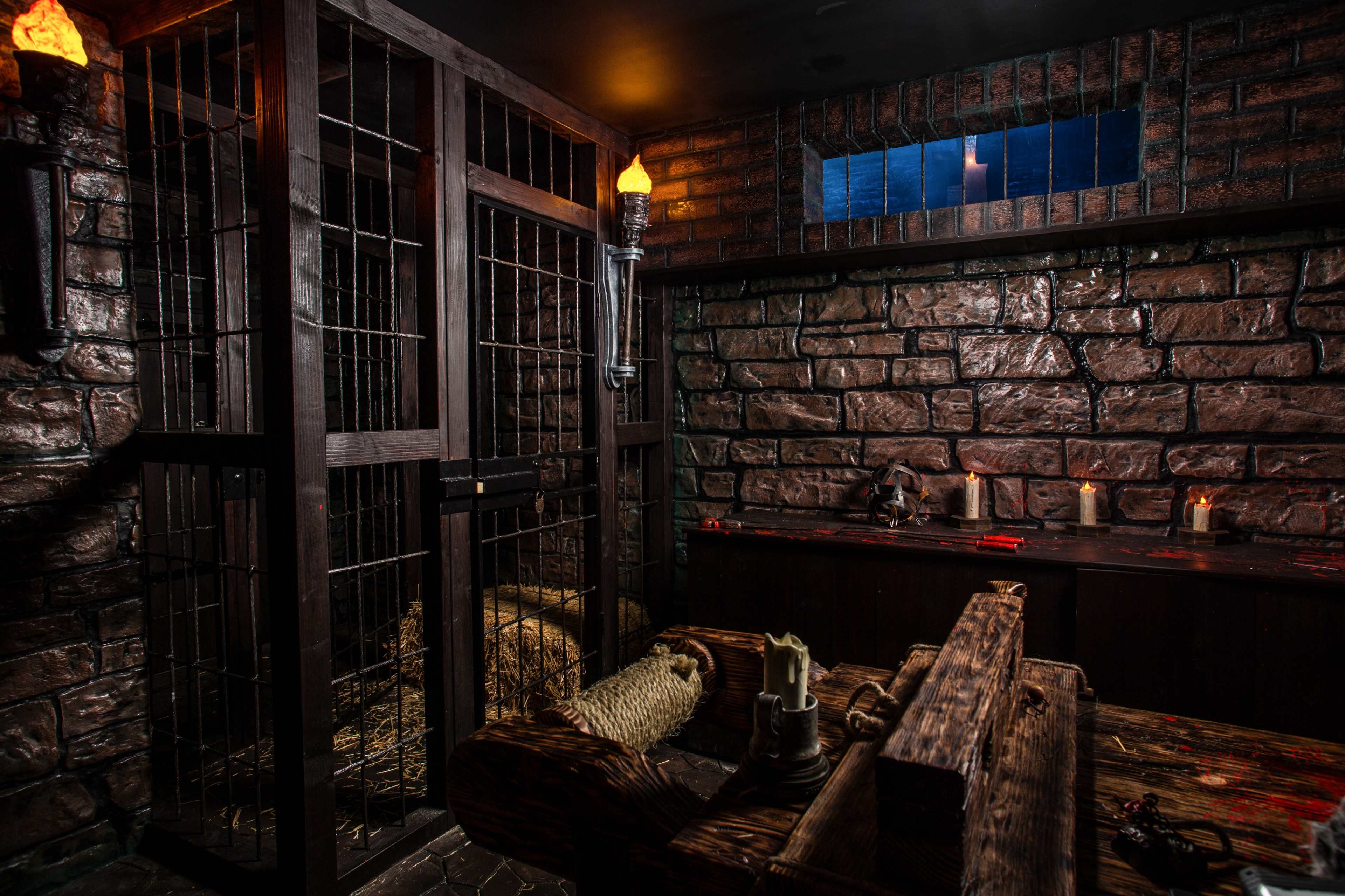 A dimly lit dungeon with stone walls, a barred cage, and a wooden stocks contraption, featuring scattered hay and candles.
