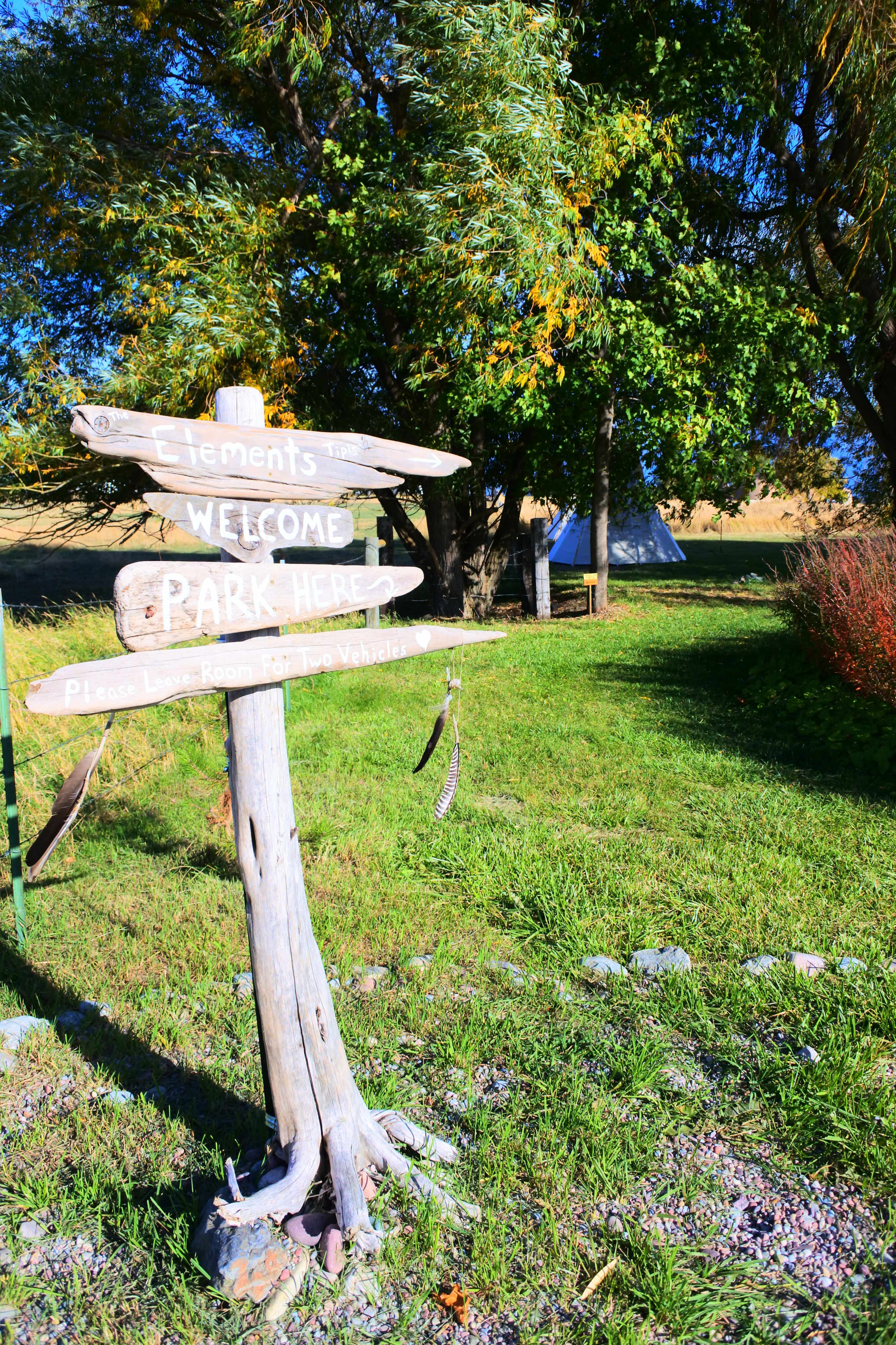 A wooden signpost with directional arrows stands in a grassy area, indicating a welcome and park entrance alongside leafy trees and a tent in the background.